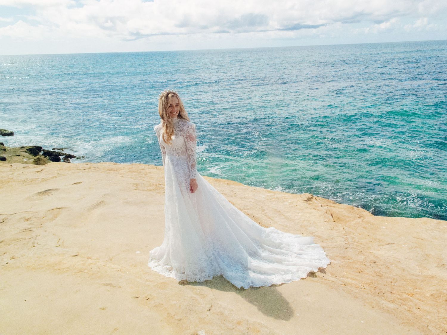Bride in elegant lace wedding gown poses on sandy cliffs overlooking turquoise ocean waters.