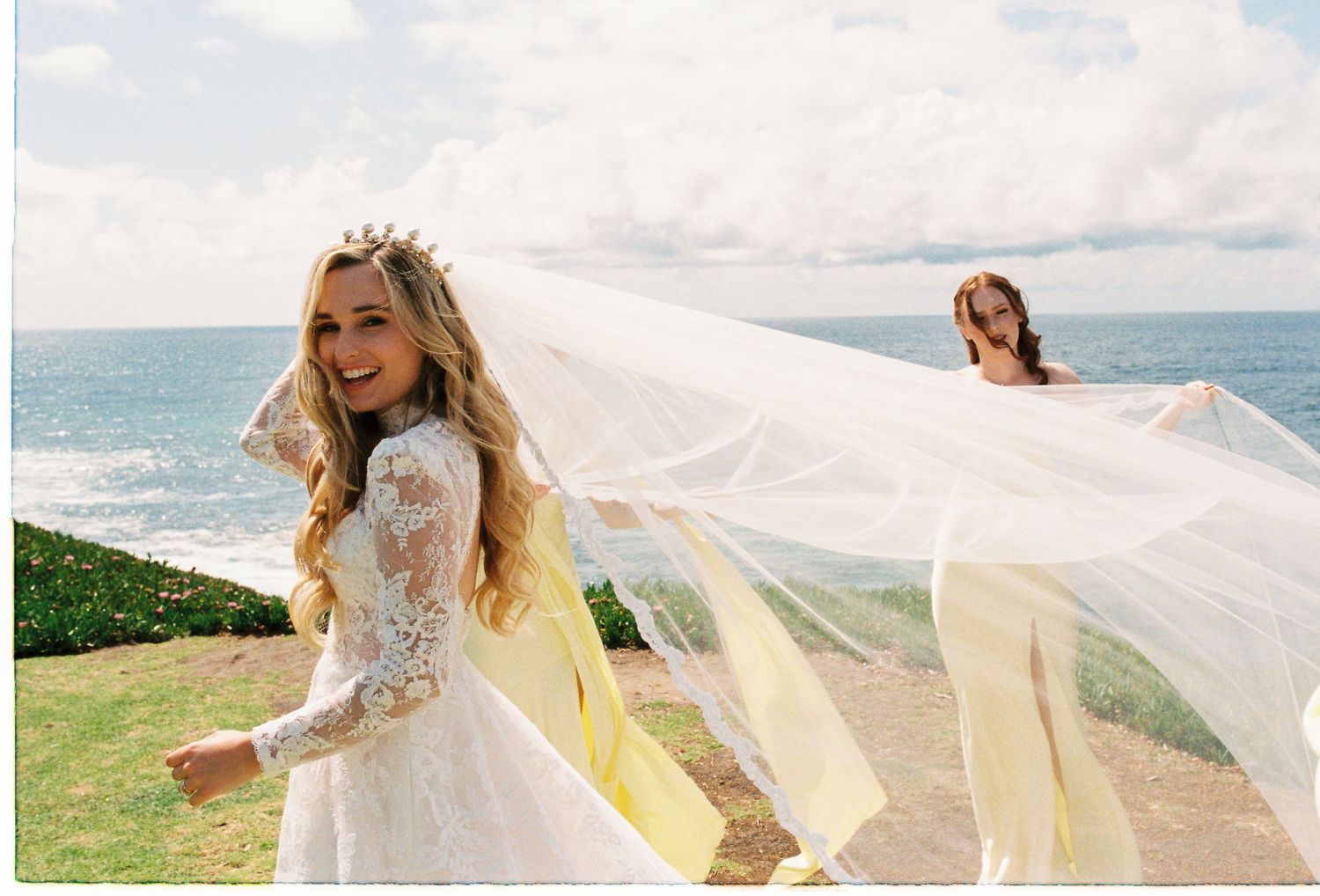 Bride in lace gown and crown smiles as her veil billows in ocean breeze with bridesmaid behind her.