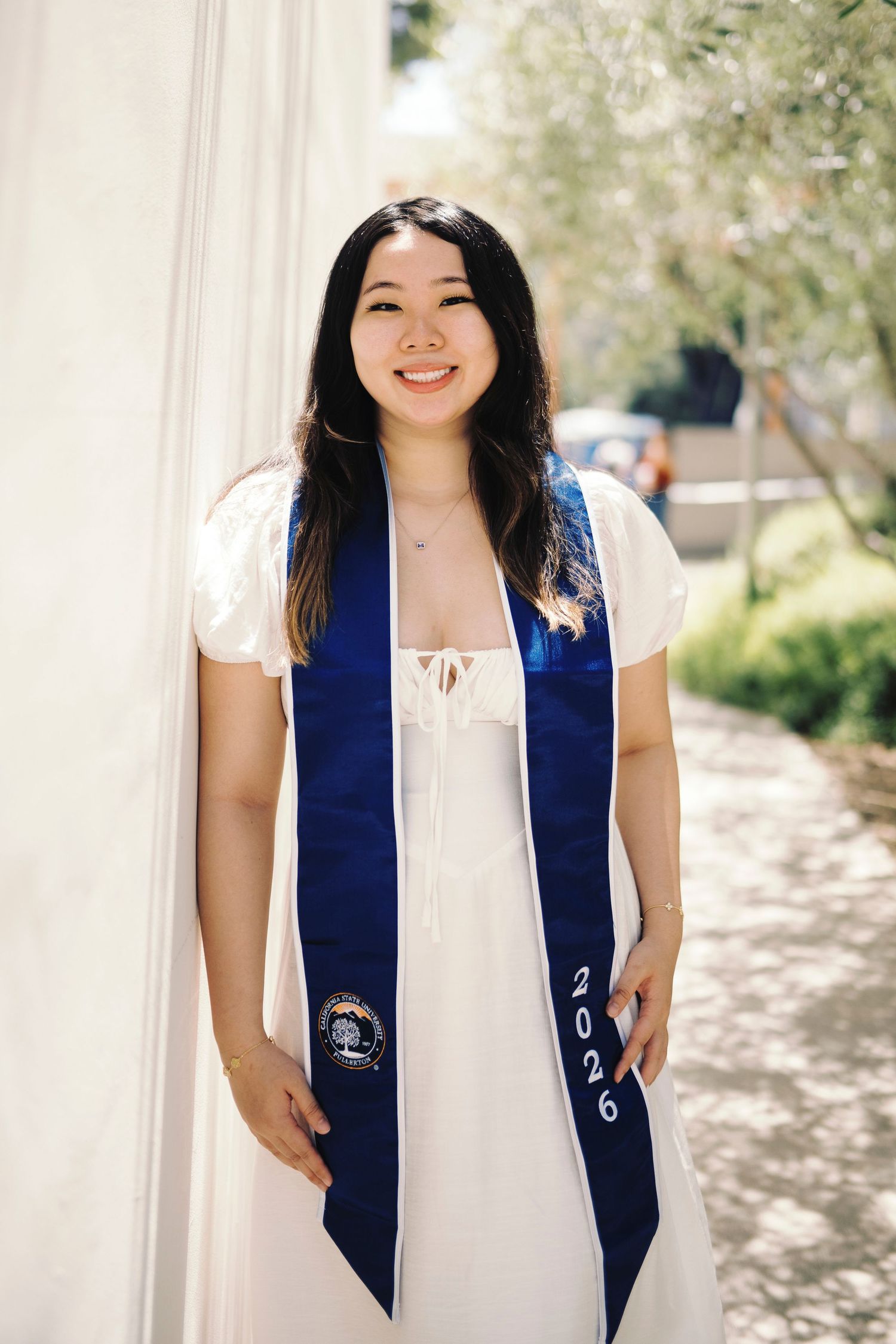 Smiling graduate wearing class of 2026 blue stole poses in white dress by white column.