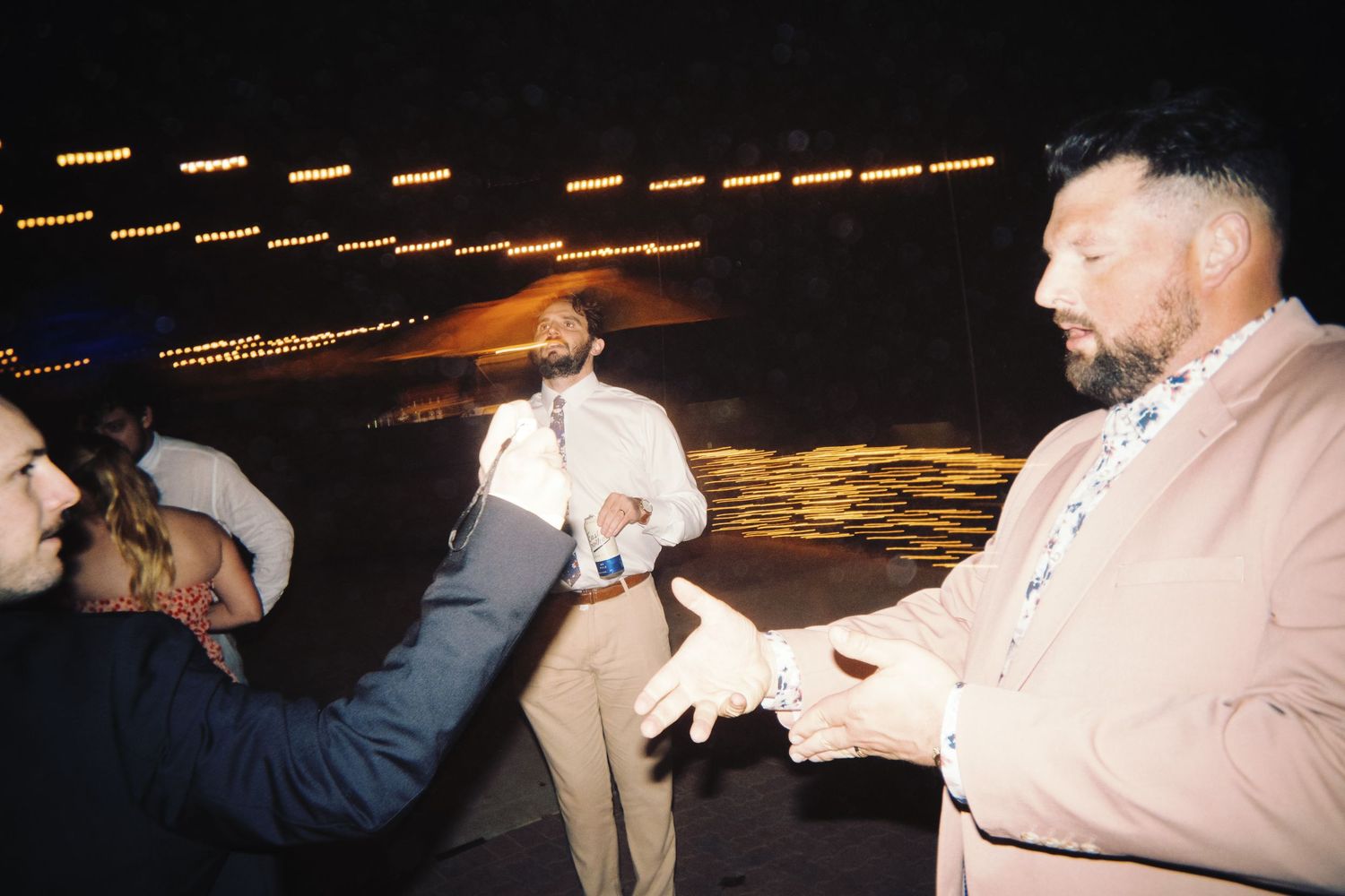 Three men in formal attire interact on outdoor wedding dance floor under glowing string lights at night.