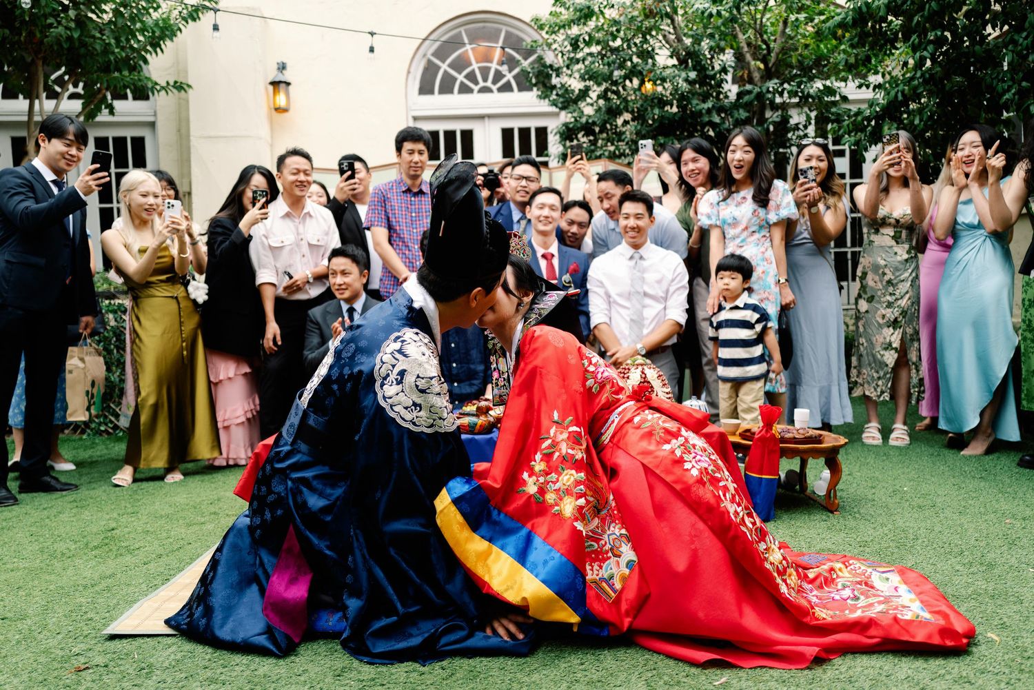 Korean wedding couple in hanbok bowing during paebaek ceremony surrounded by celebrating guests.