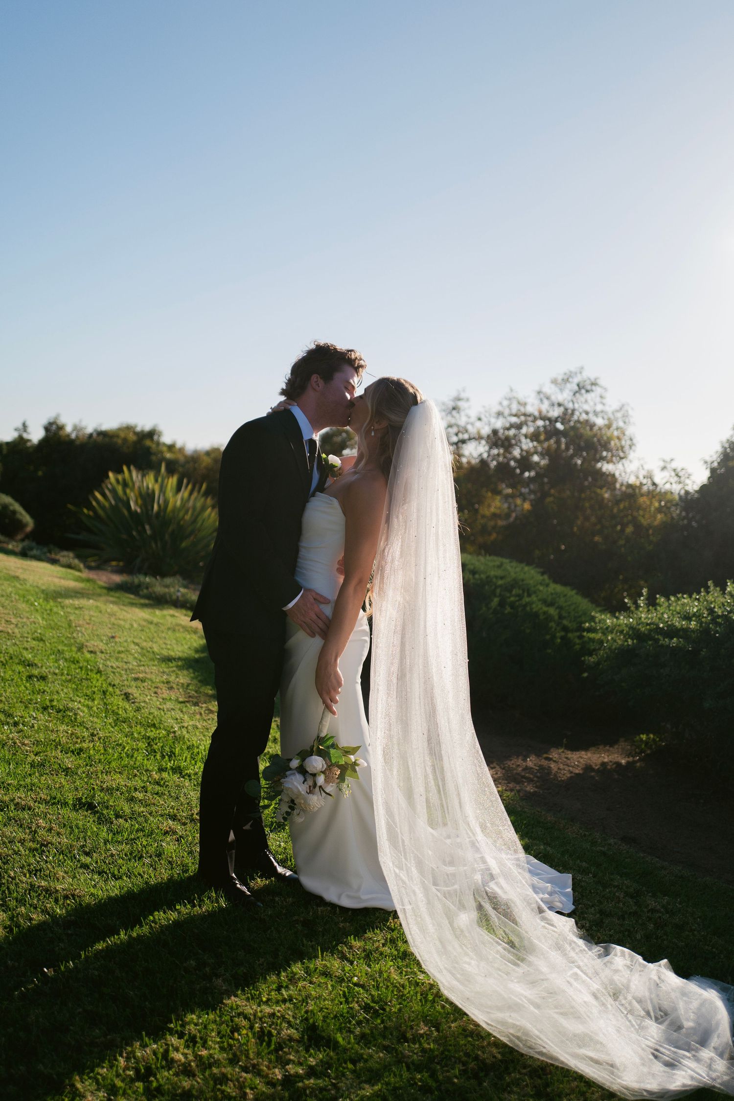 Bride and groom sharing a romantic kiss on a green lawn with a long flowing veil and bridal bouquet.