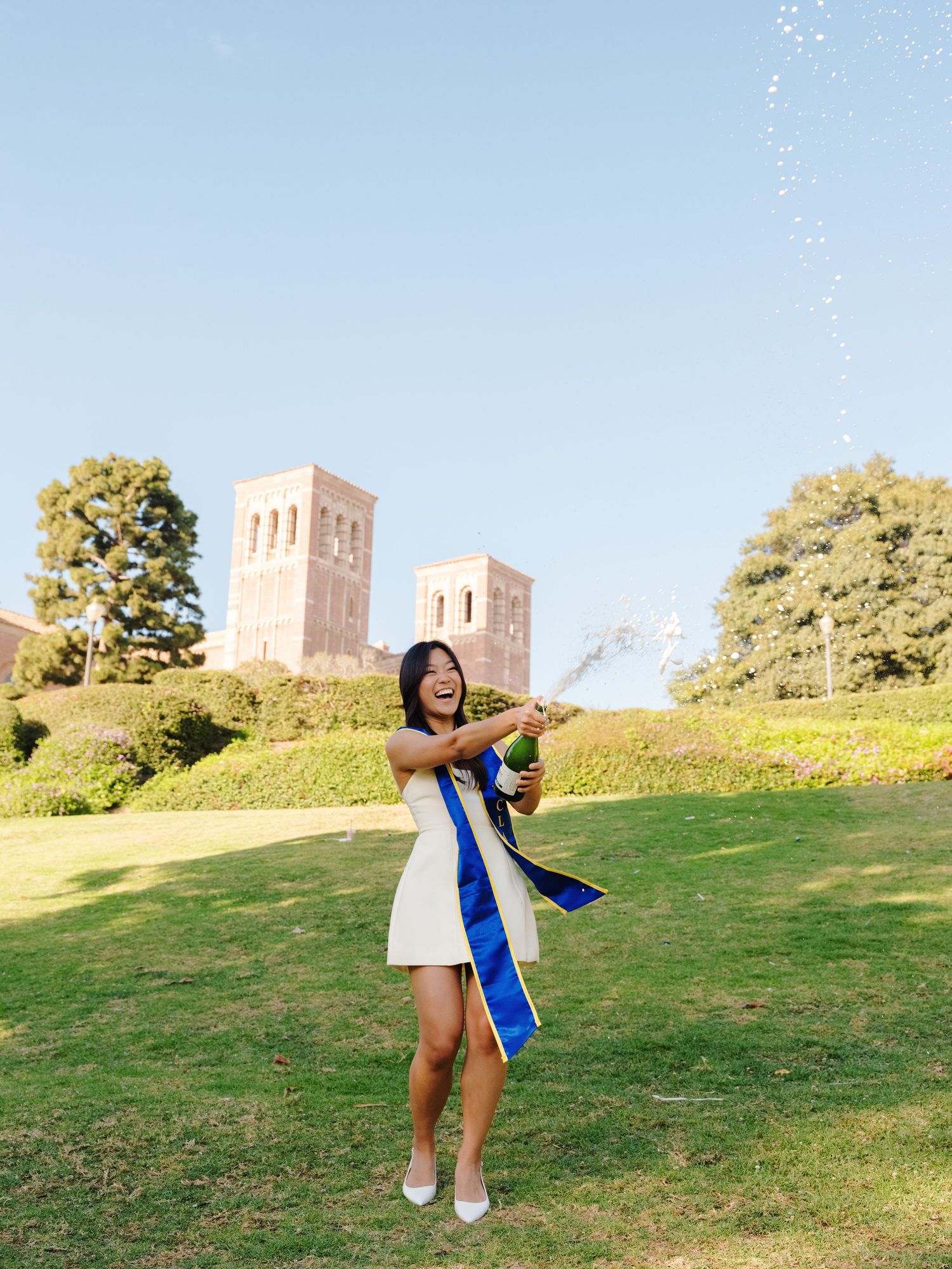 Graduate in white dress pops champagne bottle on UCLA campus lawn with towers behind her.