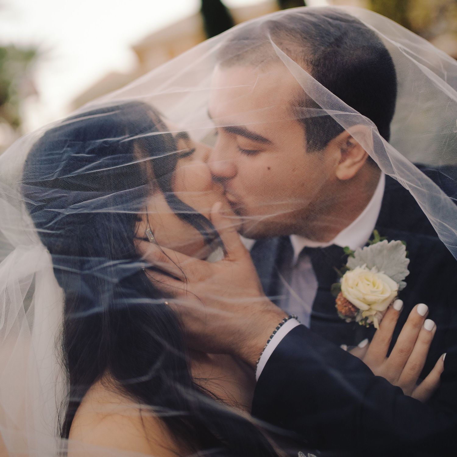 Bride and groom sharing a romantic kiss under a flowing white wedding veil outdoors.