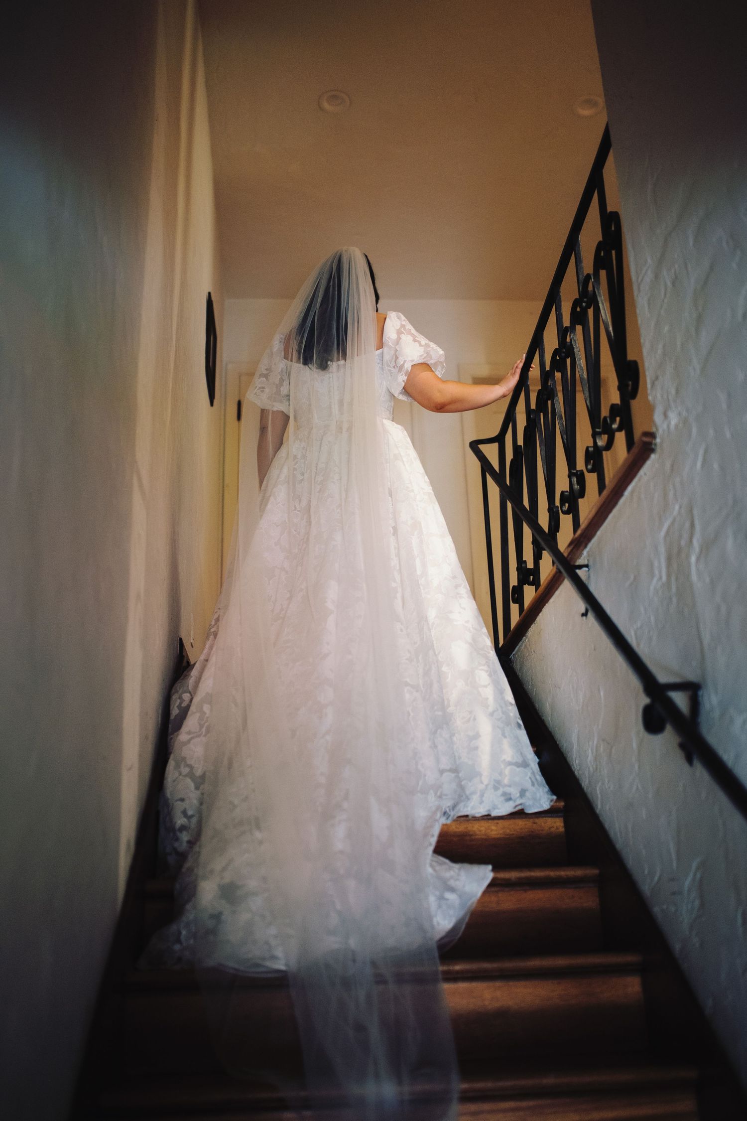 Bride in white lace ball gown and veil ascending wooden staircase with wrought iron railing.