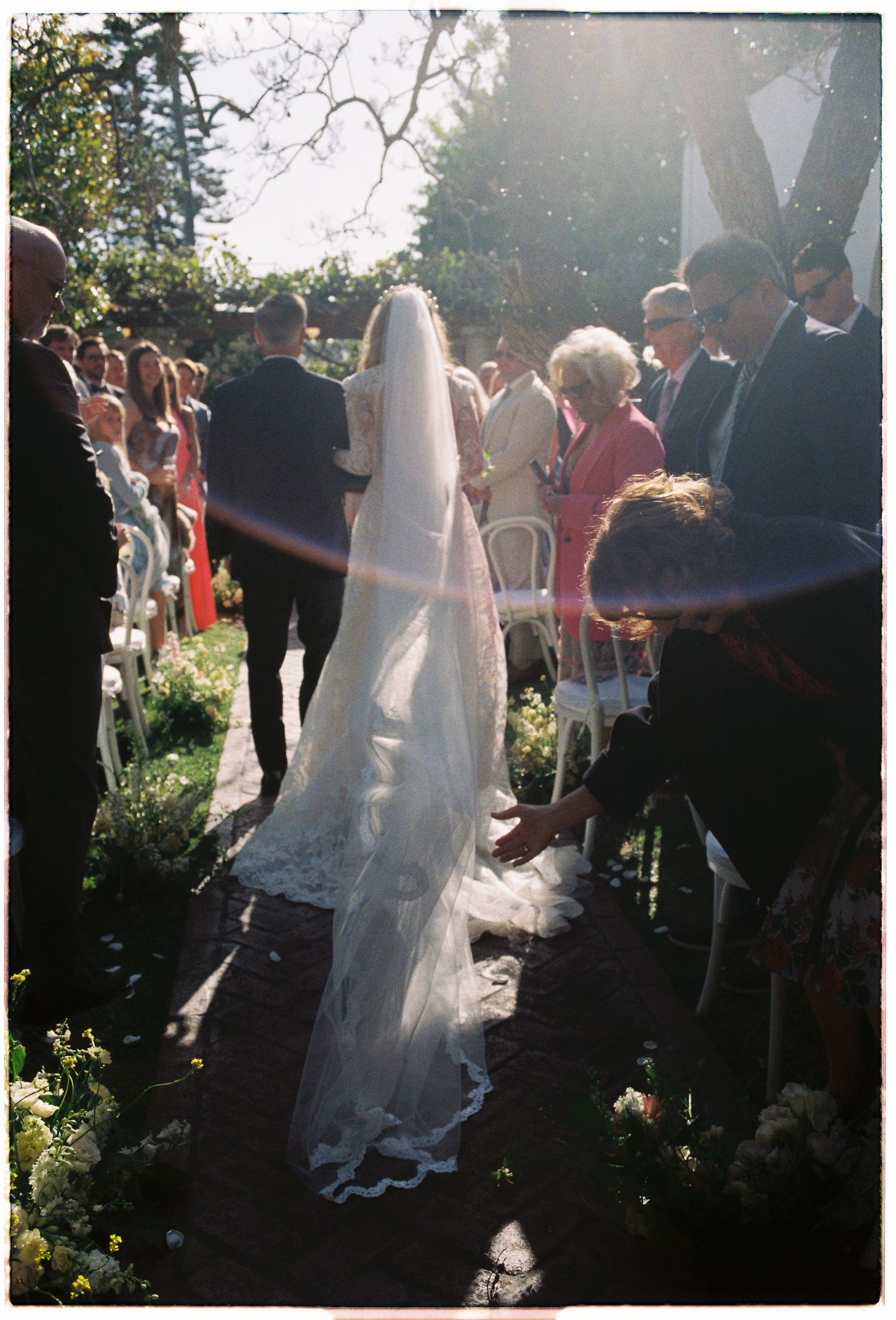 Bride in lace gown with long trailing veil walks down sunlit garden aisle surrounded by guests.