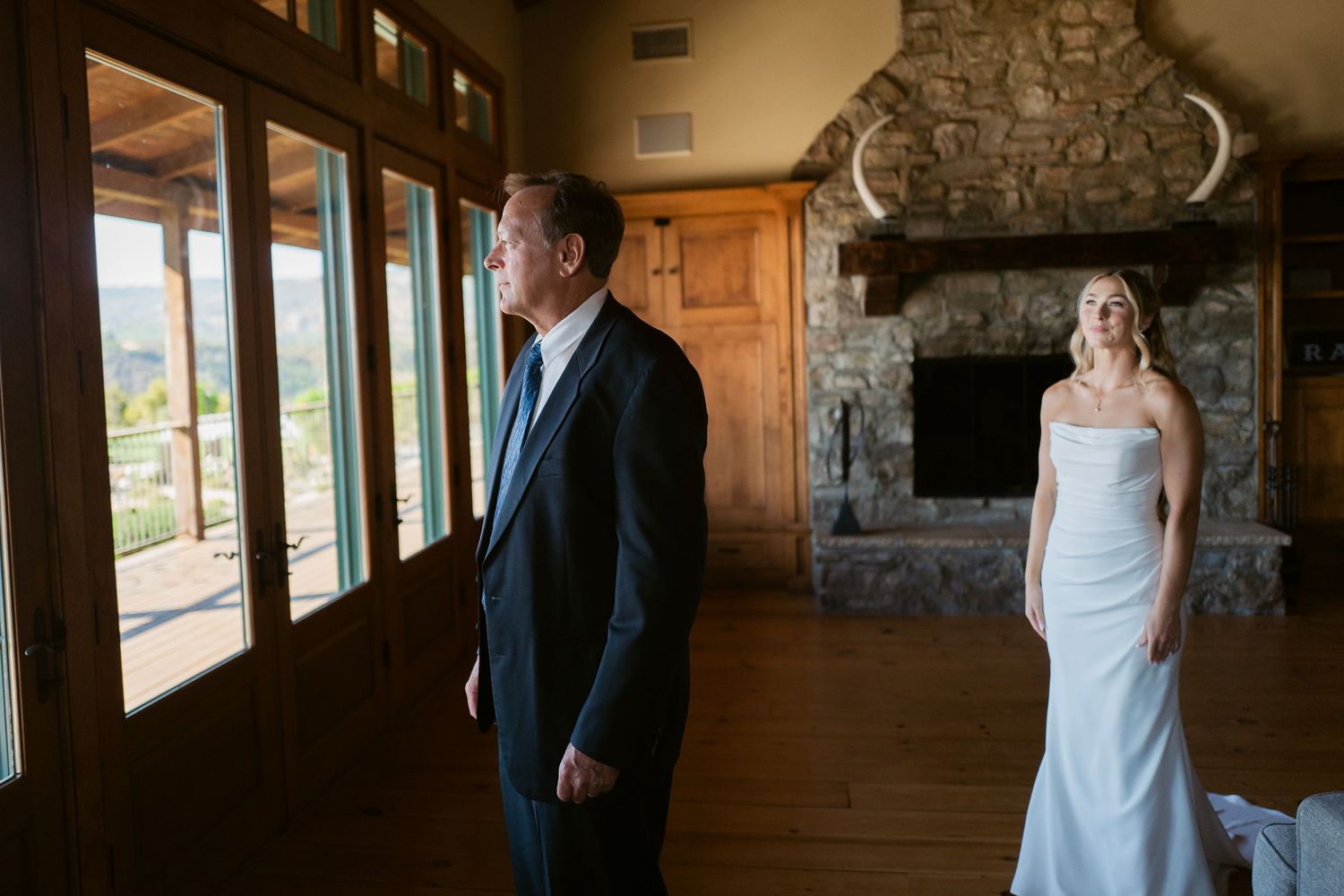Bride in white gown approaches groom in suit near stone fireplace during wedding first look.