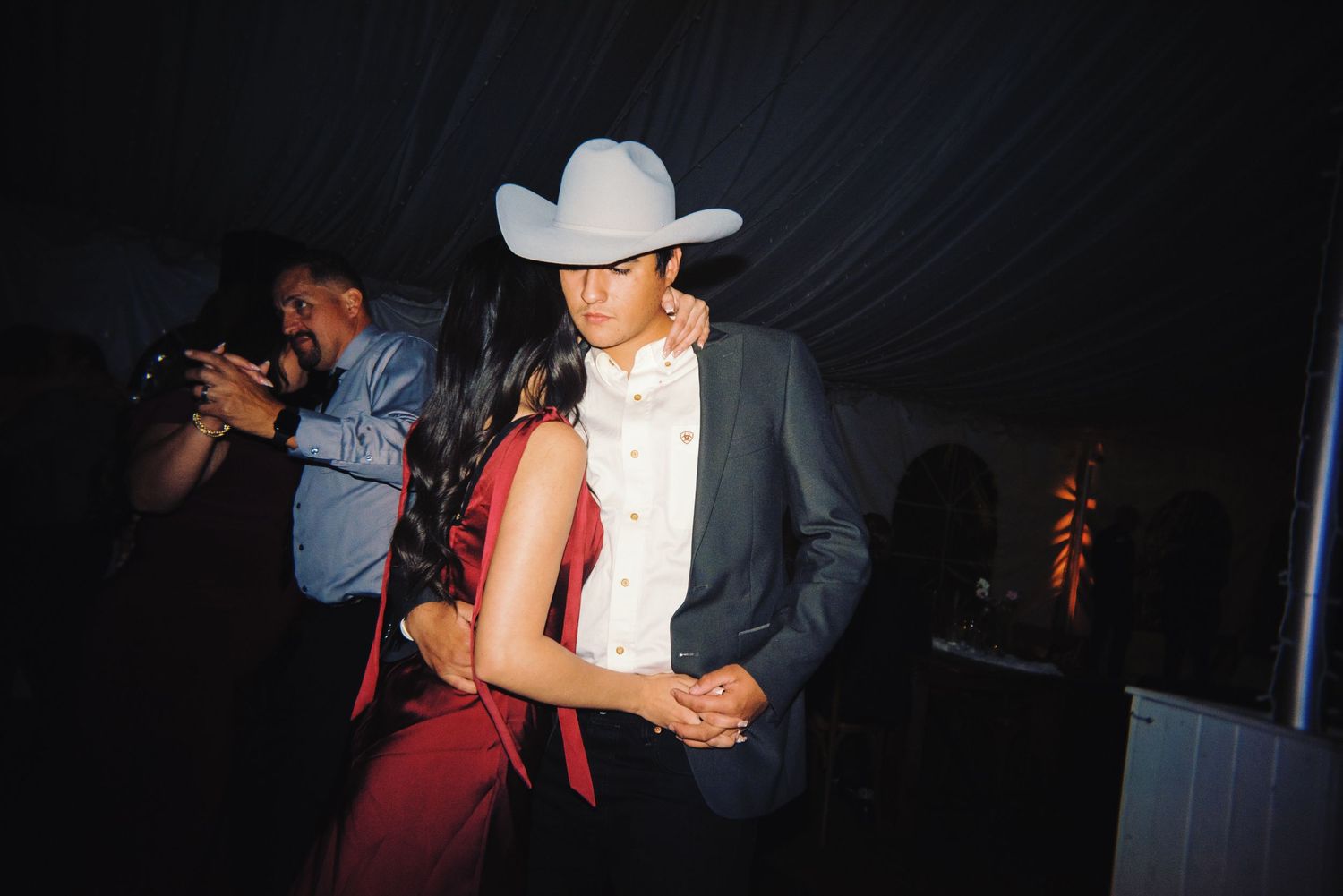Couple dancing closely at wedding reception with guest wearing white cowboy hat.