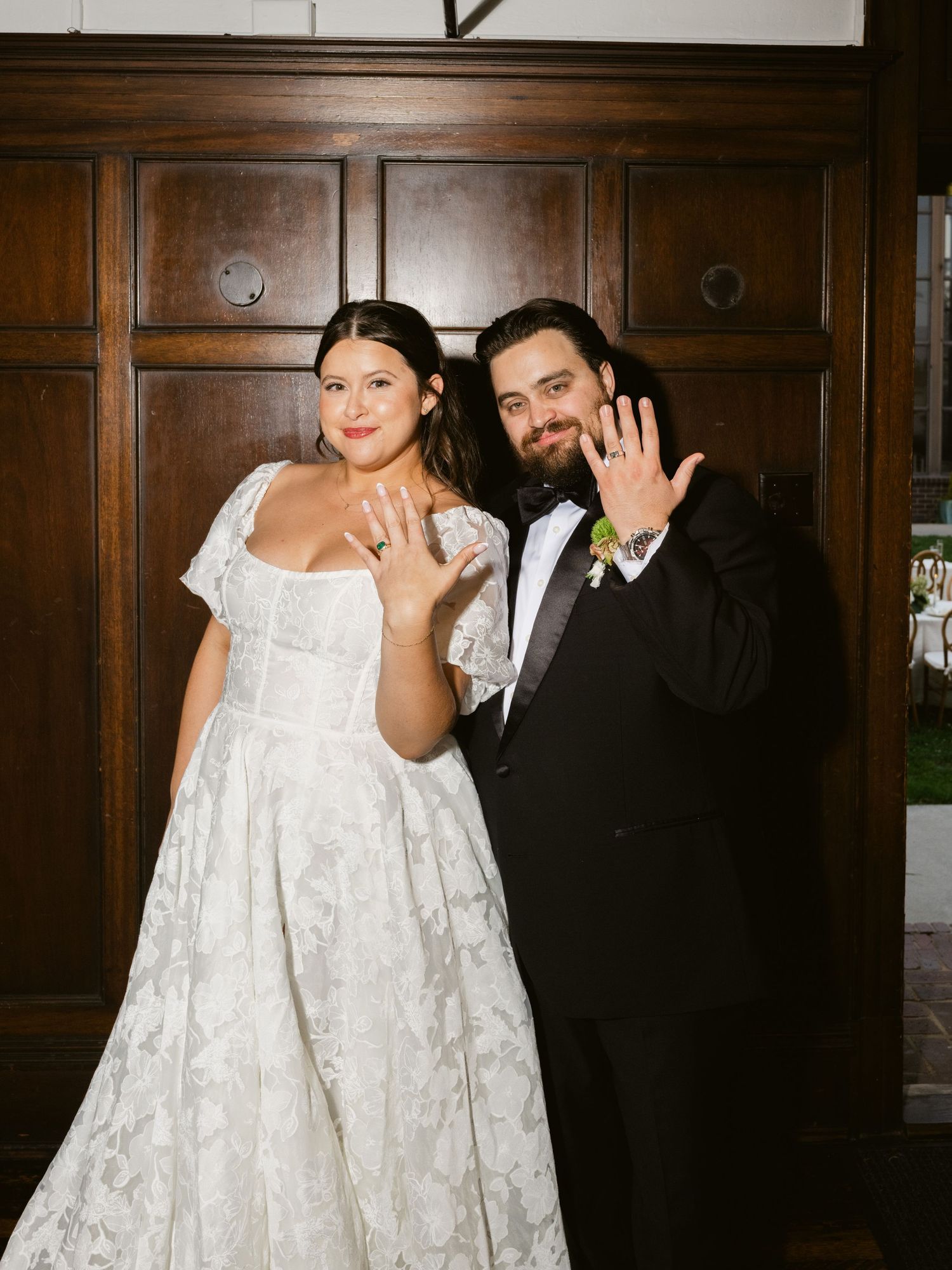 Newlyweds show off wedding rings while posing in front of wooden doors indoors.
