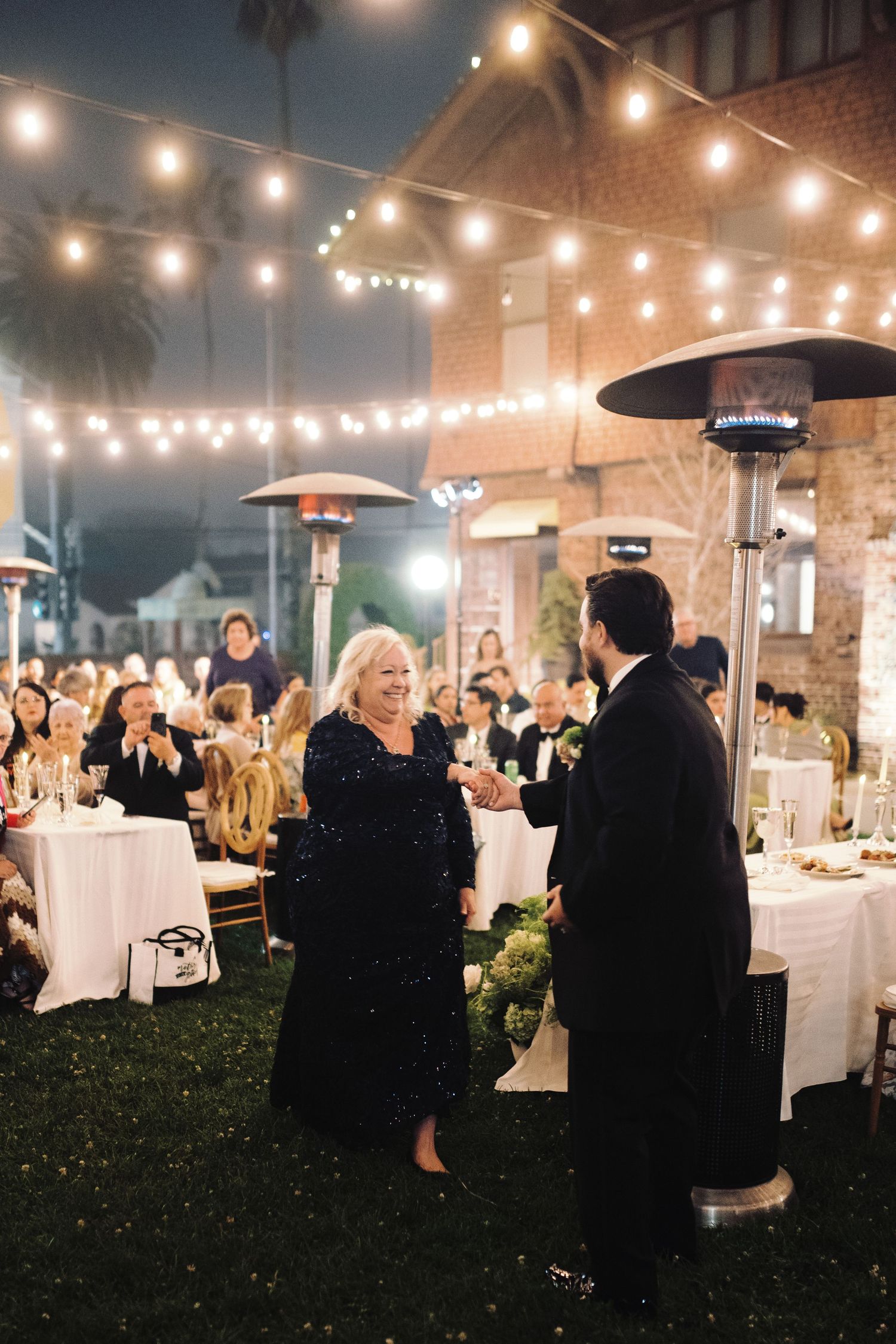 Groom dances with woman in navy sequin dress under string lights at outdoor wedding reception.