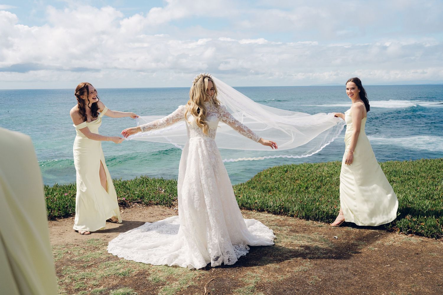 Bride in lace gown extends her veil as two bridesmaids hold it near a stunning ocean overlook.