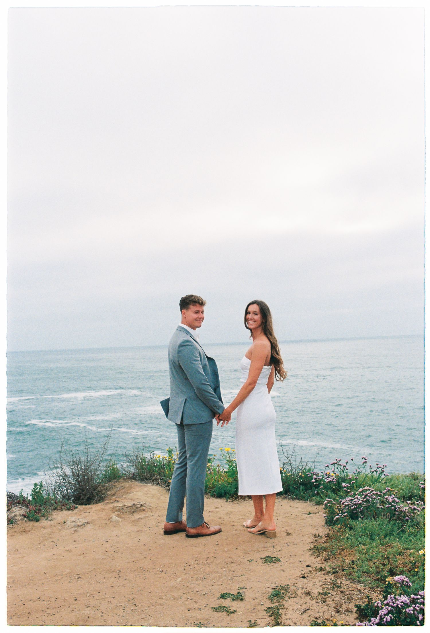 Engaged couple holding hands on coastal cliff overlooking ocean, woman in white backless dress and man in gray suit smiling.
