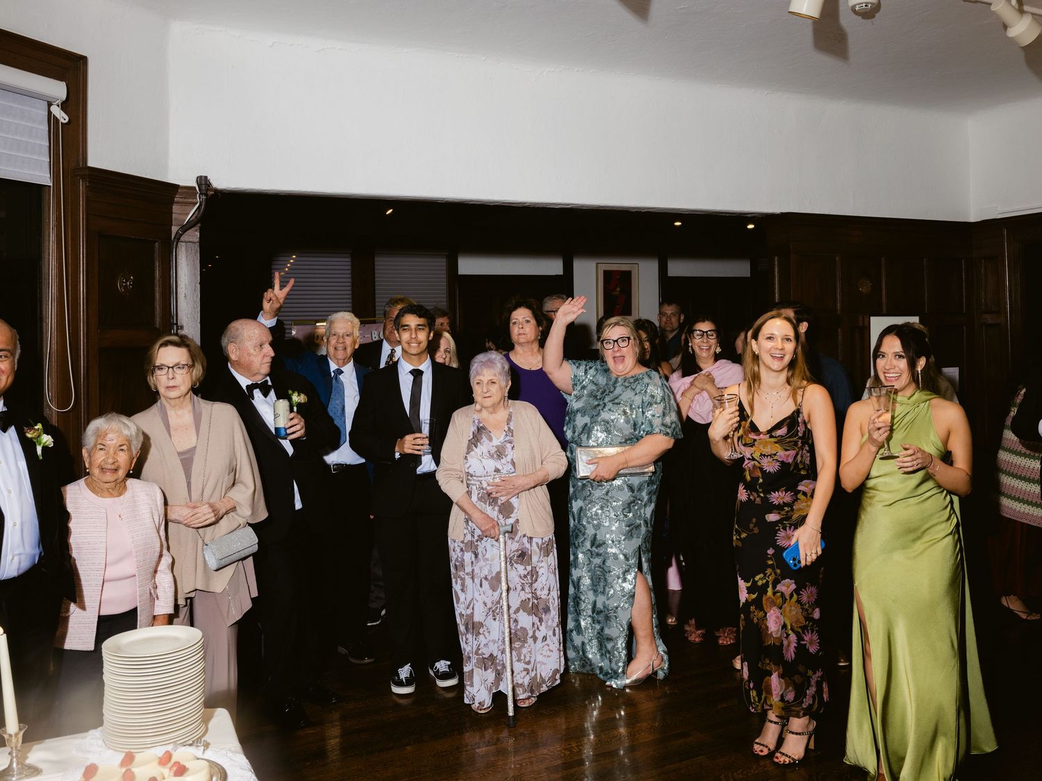 Joyful wedding guests clapping and celebrating near a tiered cake inside an elegant reception hall.