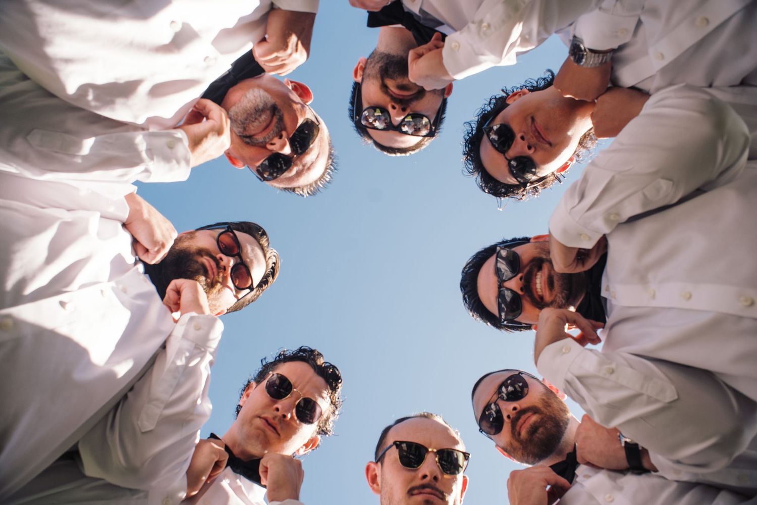 Groomsmen in white shirts and sunglasses forming circle viewed from below against blue sky.