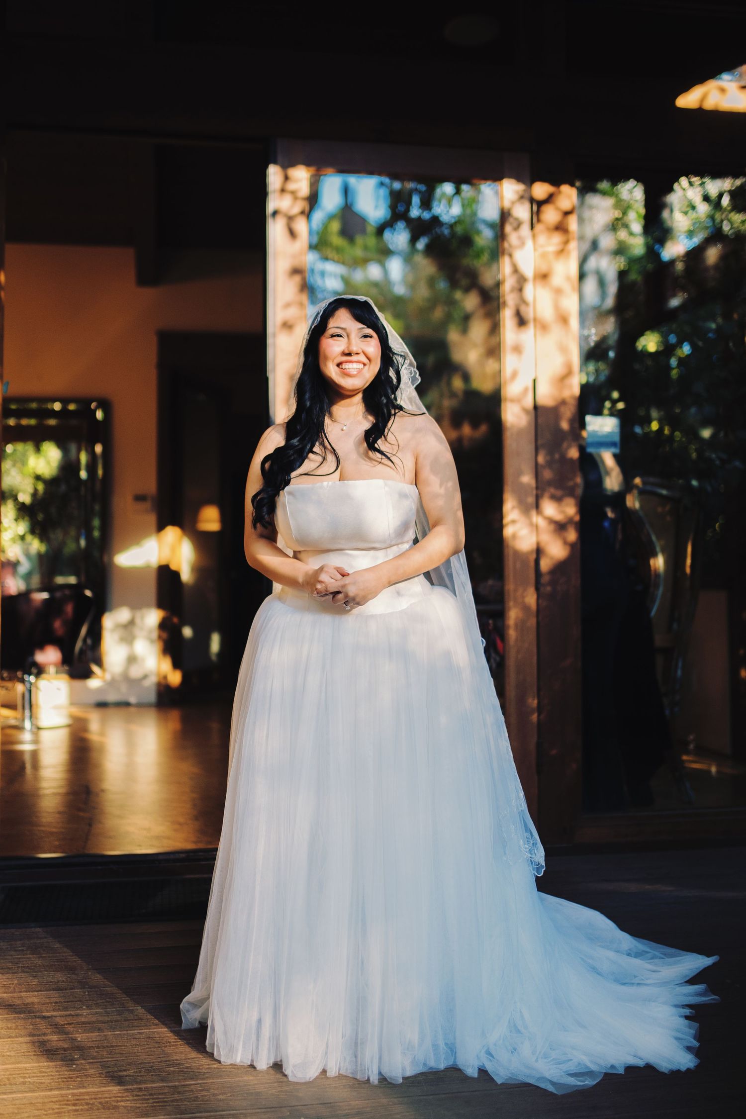 Radiant bride in strapless tulle ball gown and veil smiles in a warmly lit wedding venue.