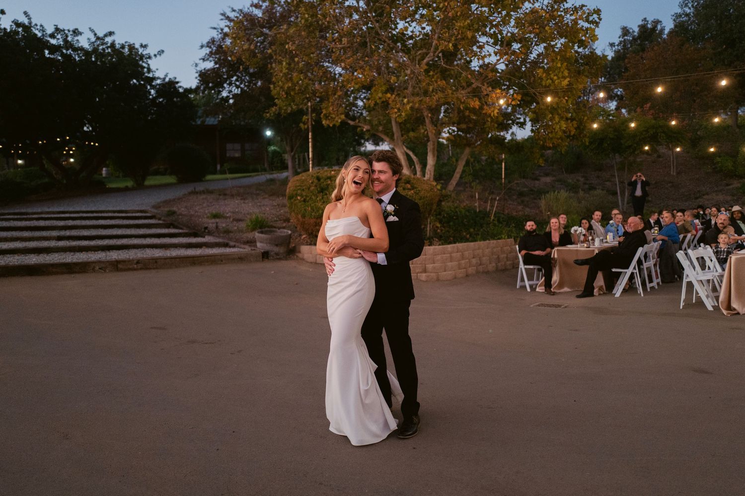 Bride and groom share a romantic first dance on an open-air patio under string lights with seated guests nearby.