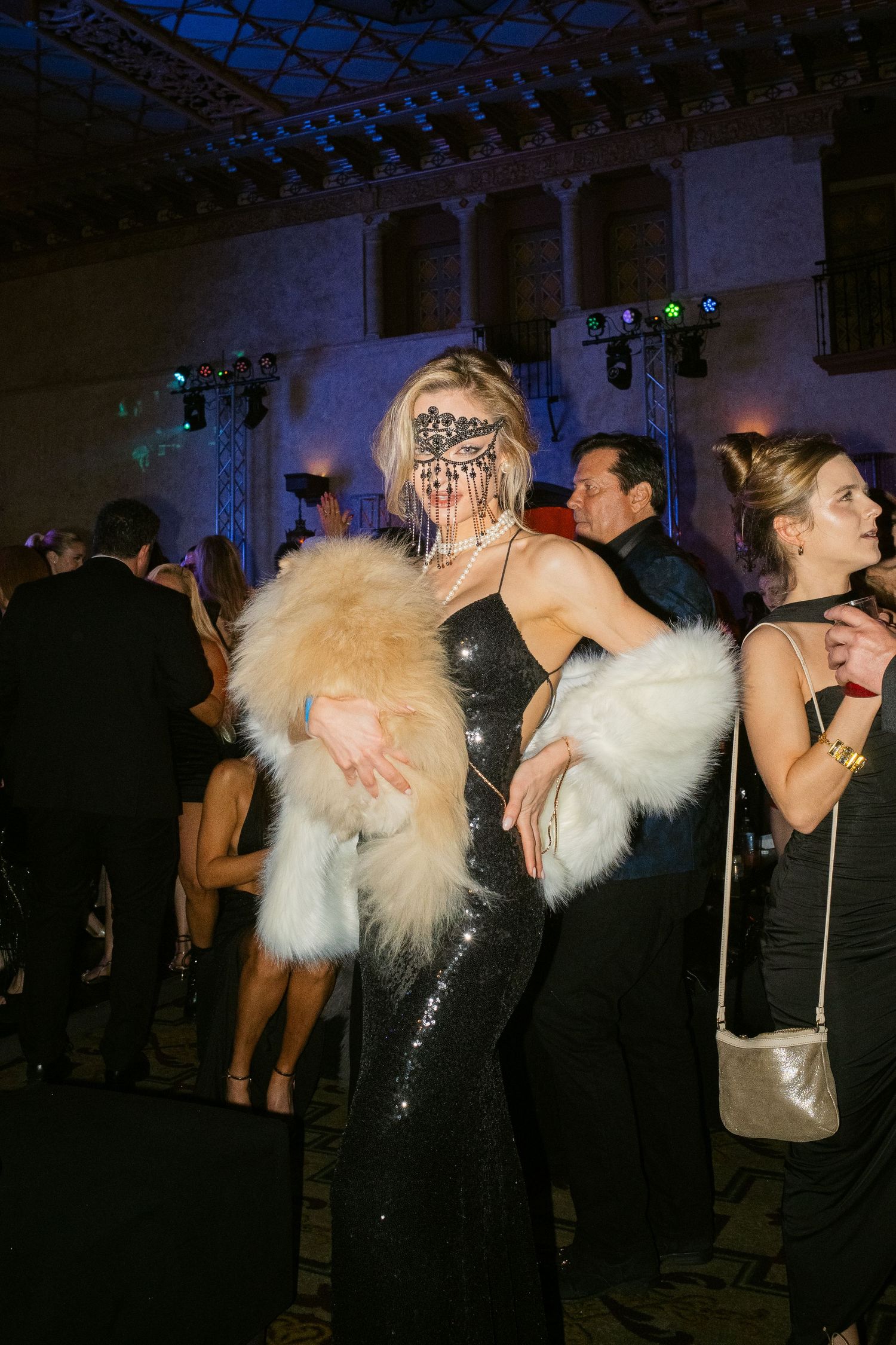 Woman in black sequin gown, lace masquerade mask, and fur stole poses at elegant masquerade ball.