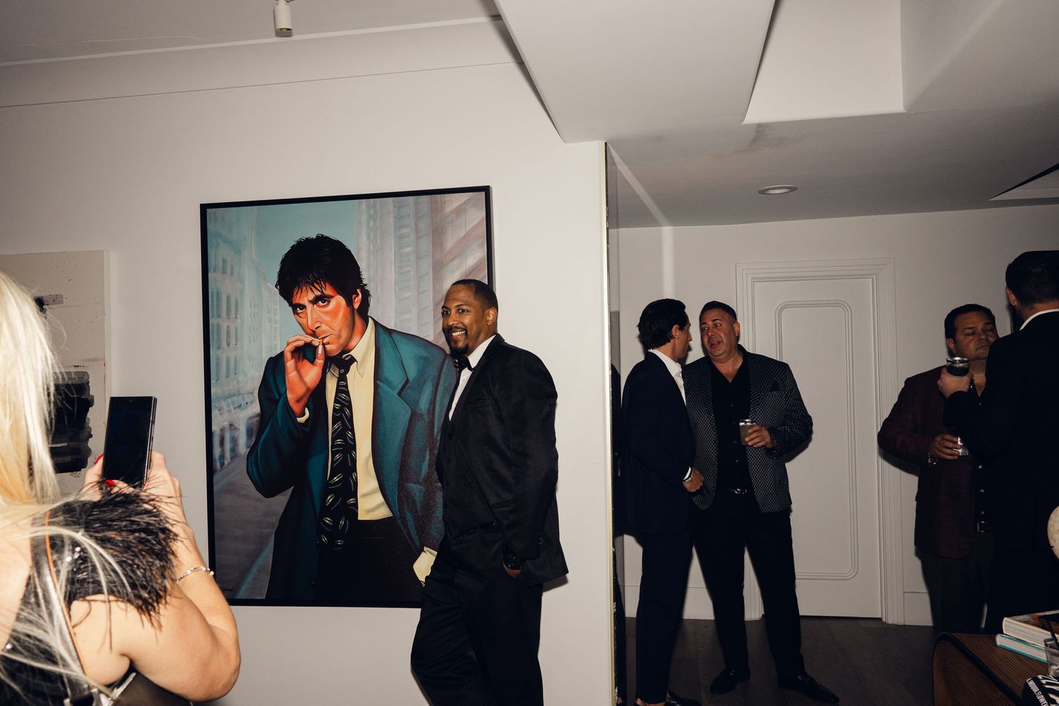 Guests mingle at an art gallery opening with a large portrait painting displayed on the wall behind them.