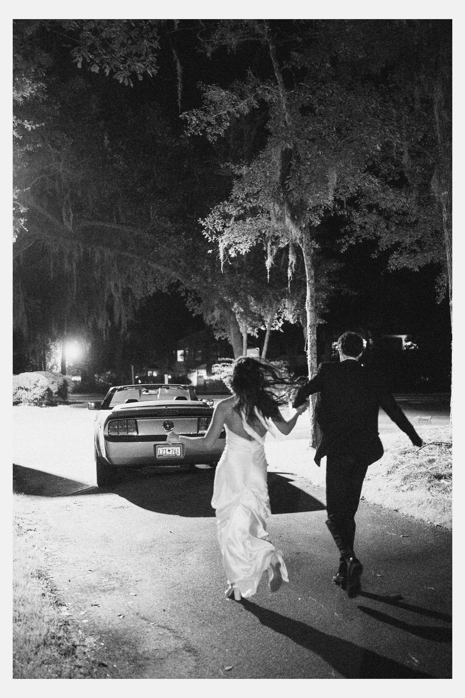 Bride and groom walking hand in hand at night toward a classic car, black and white wedding photography.