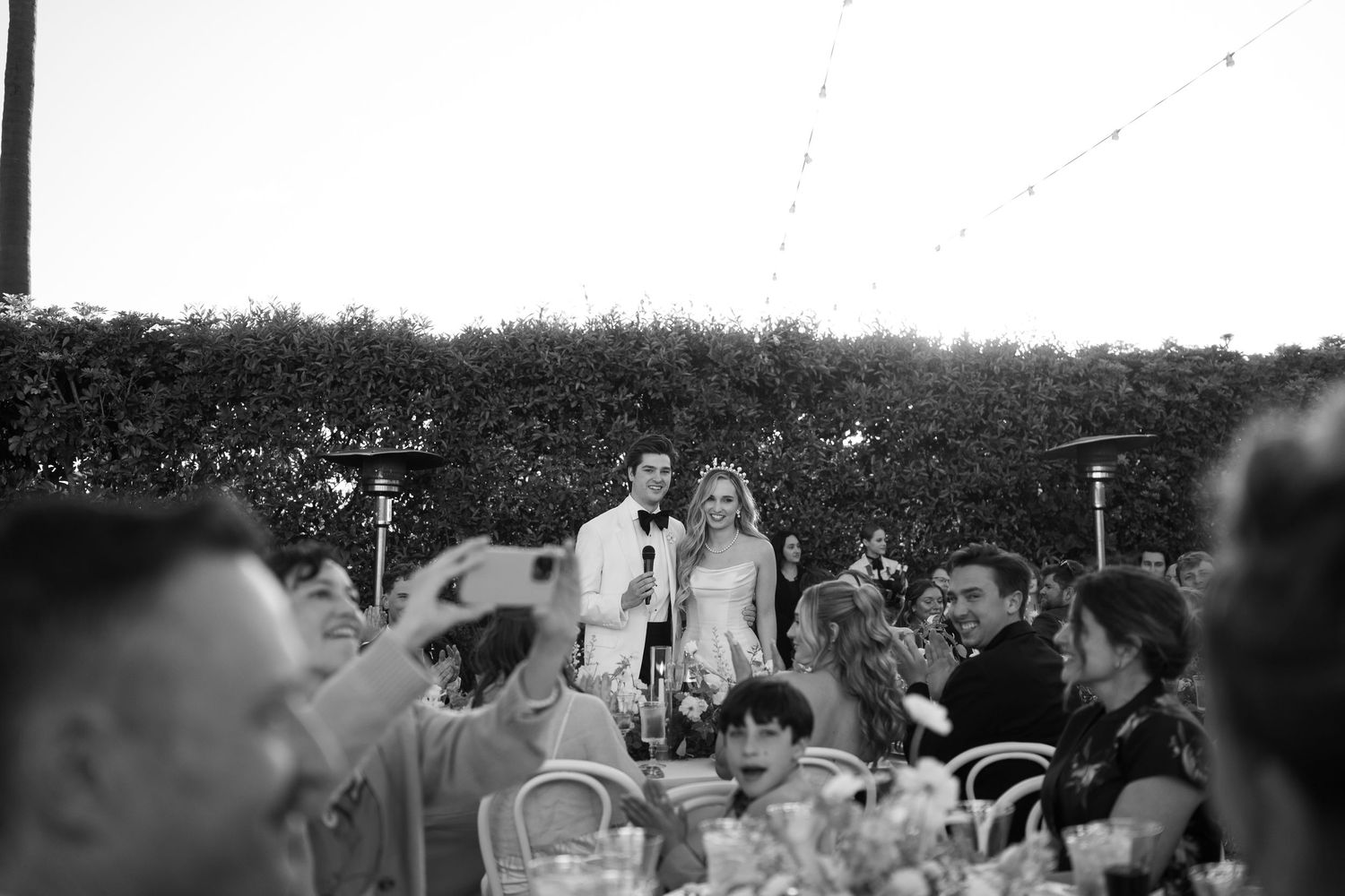 Bride and groom stand at reception table during wedding speeches under string lights in black and white.