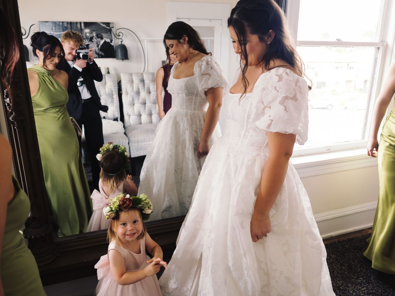 Bride in lace gown smiles at adorable flower girl in floral crown with bridesmaids in green dresses.