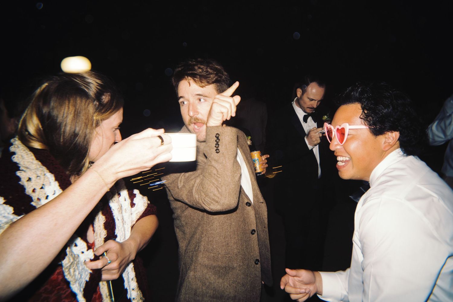 Guests laughing and socializing at a lively evening party, one wearing heart-shaped glasses.
