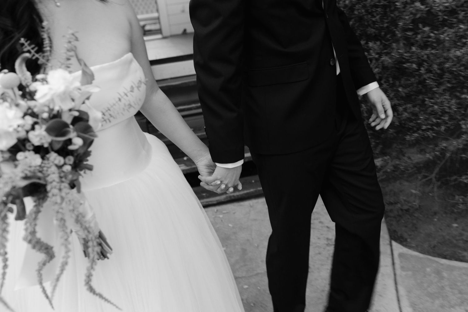 Black and white photo of bride and groom holding hands, bride carrying a cascading wedding bouquet.