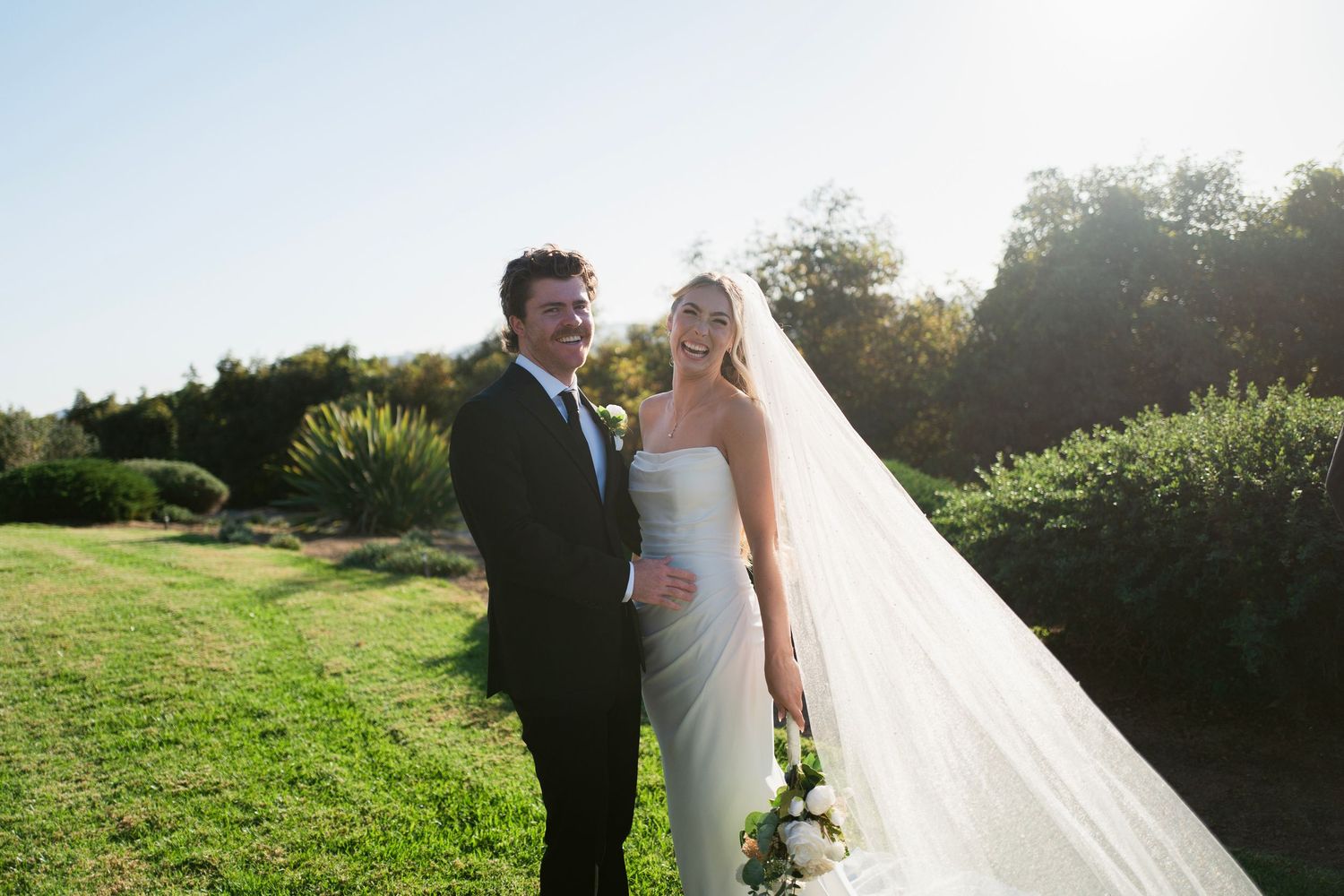 Smiling bride in strapless gown with cathedral veil stands beside groom in black suit outdoors.