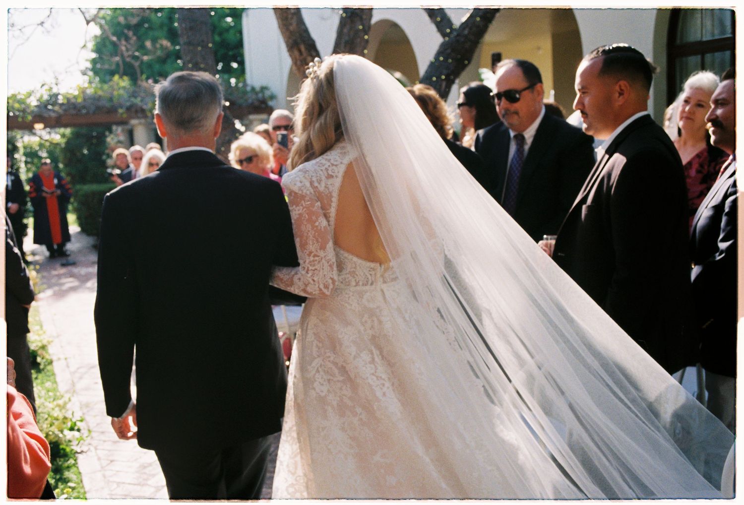 Bride in lace gown with long veil walks down aisle escorted by father at outdoor wedding ceremony.