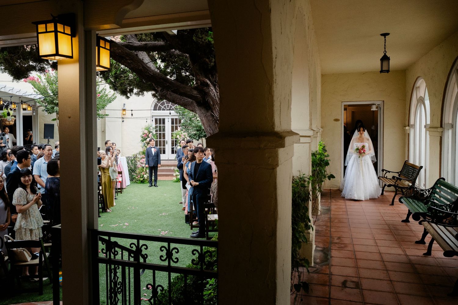Bride in a flowing gown walking along a Spanish-style colonnade toward her outdoor wedding ceremony.