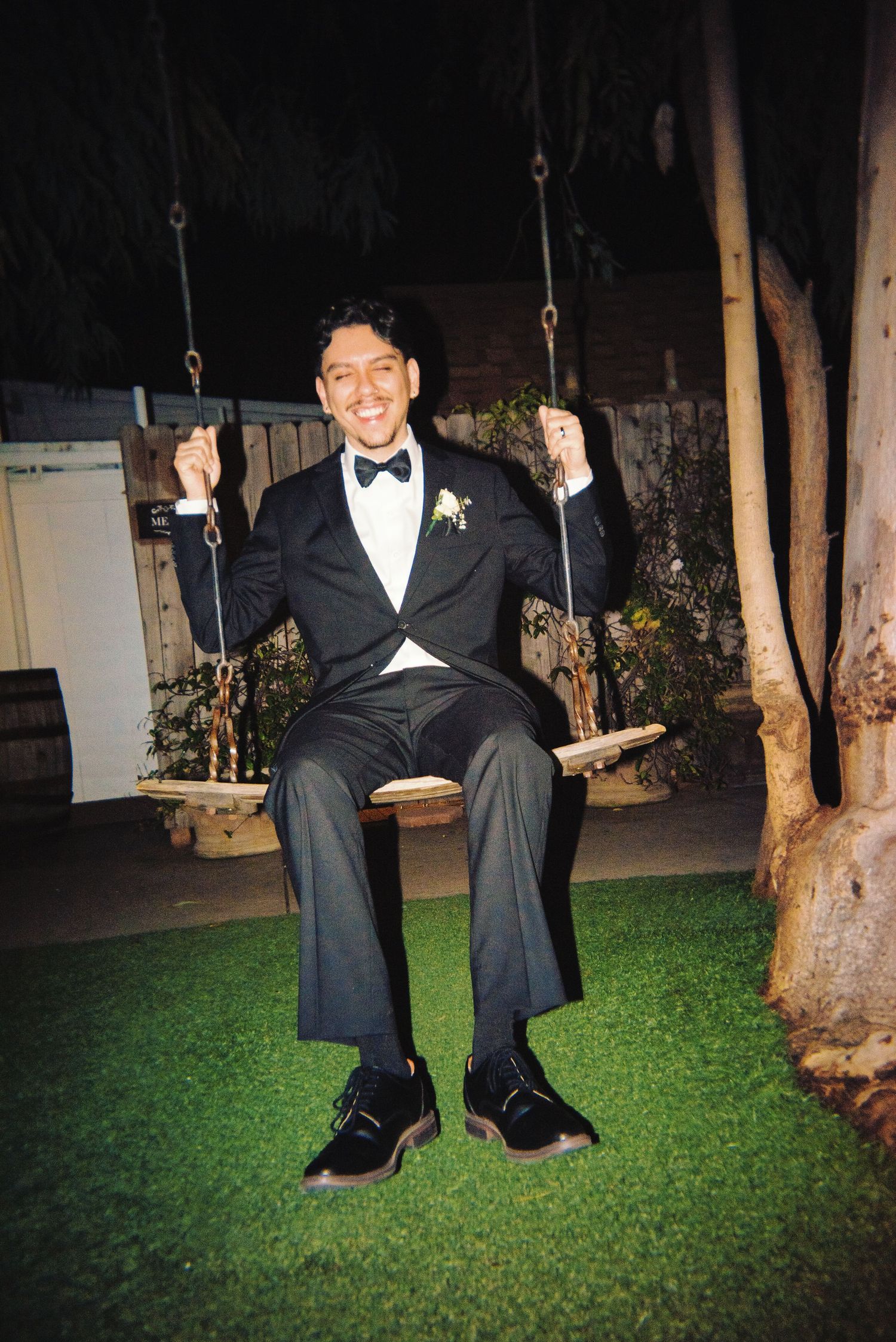 Smiling groom in tuxedo sitting on outdoor swing at nighttime wedding venue.