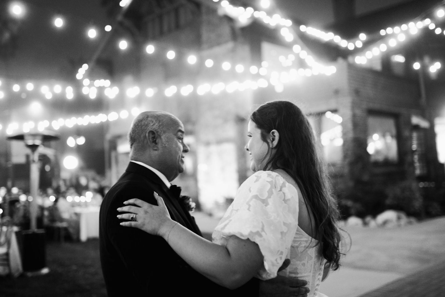 Black and white photo of bride and partner sharing intimate dance under string lights at night.