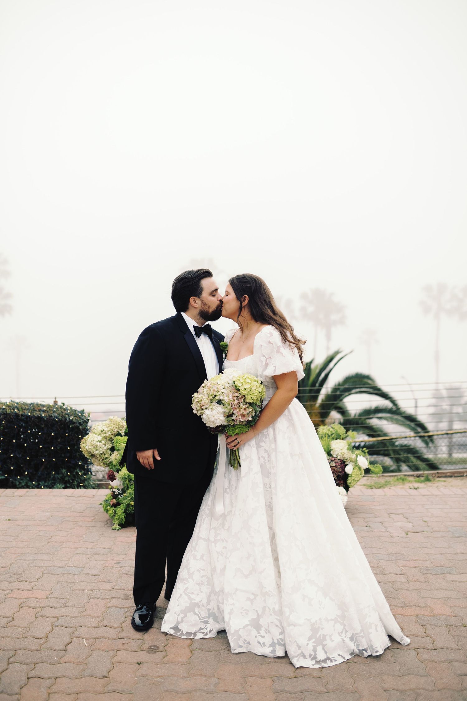 Bride and groom share romantic kiss outdoors on foggy day with palm trees.