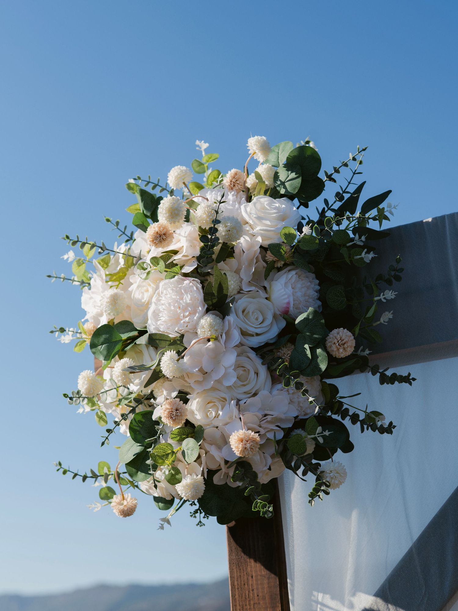 Lush white floral arrangement of peonies, roses, and eucalyptus adorning a wooden wedding arch against a blue sky.