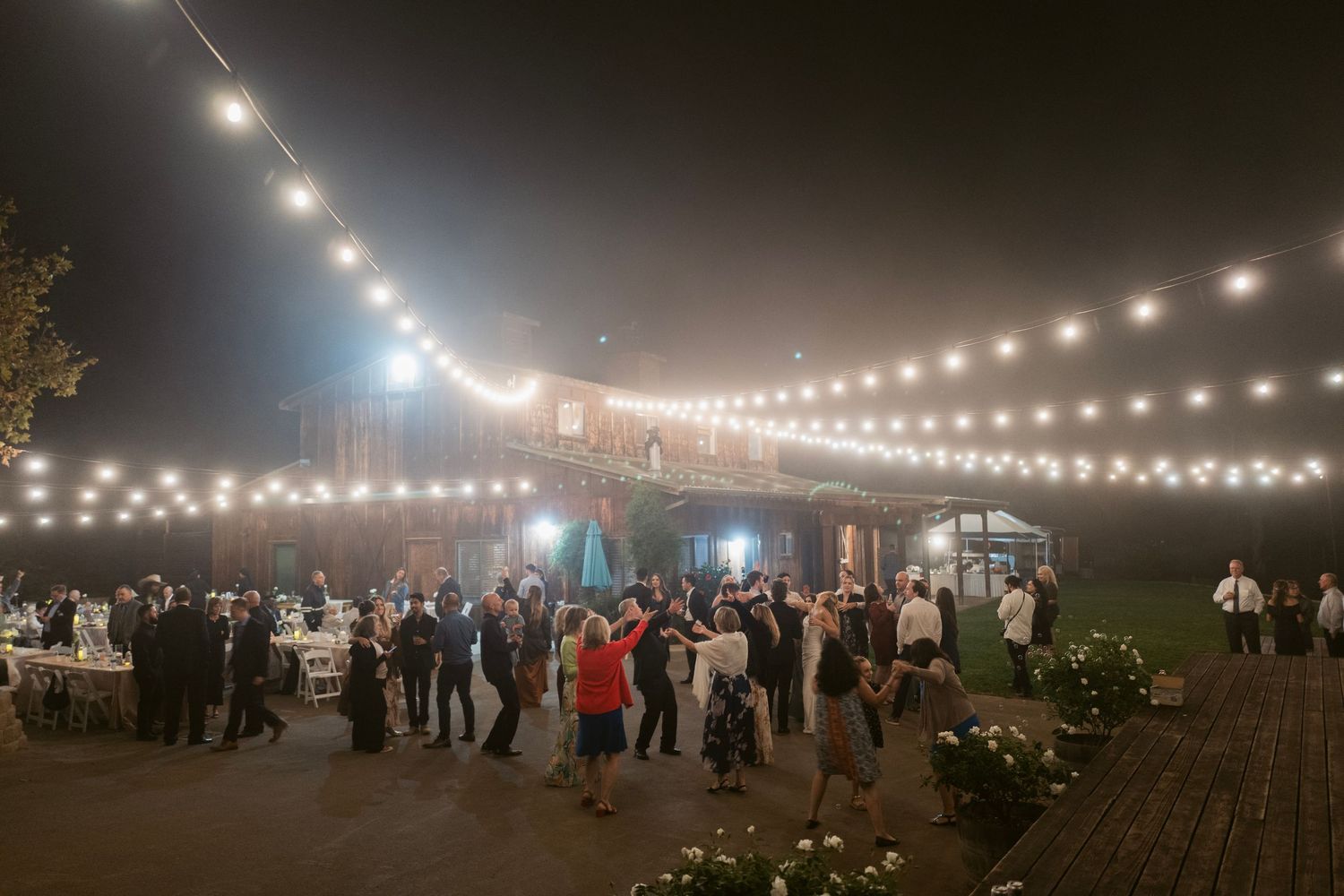 Wide shot of elegant outdoor barn wedding reception with string lights and guests mingling at night.