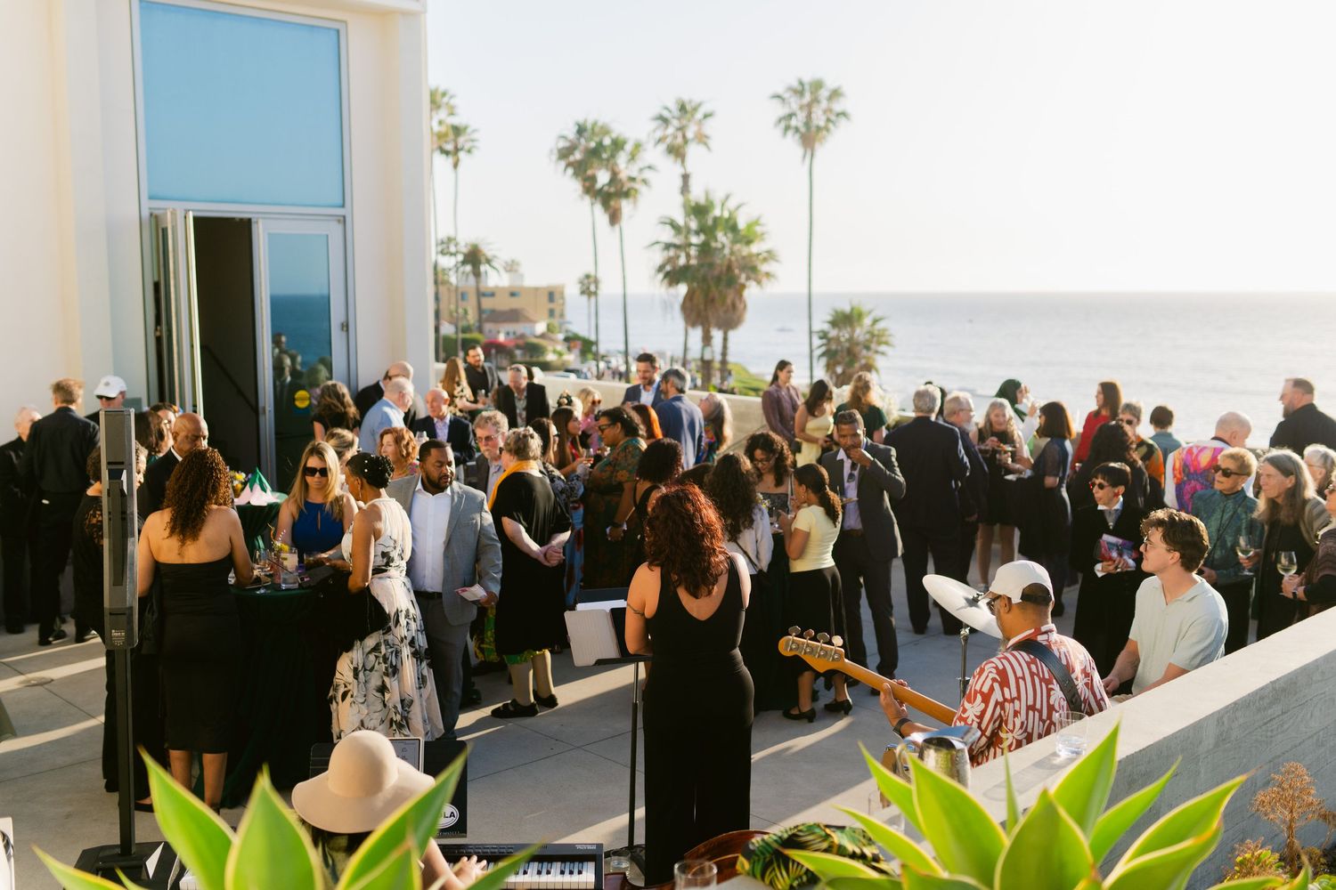 Outdoor wedding cocktail hour on ocean-view terrace with palm trees, guests mingling, and live band performing near the coast.