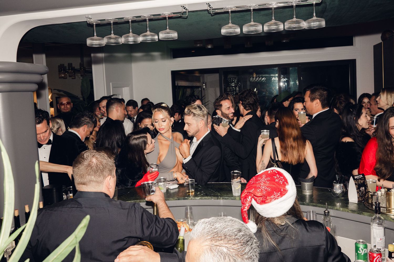 Elegant holiday party at a bar with guests in formal attire, featuring a Santa hat and hanging wine glasses.