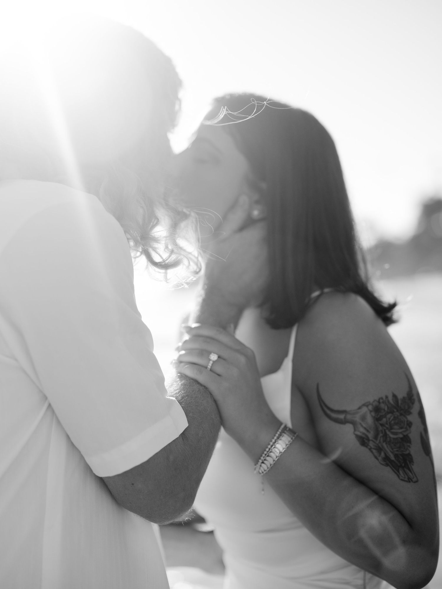 Black and white romantic photo of couple kissing in bright sunlight, woman showing engagement ring and tattoo.