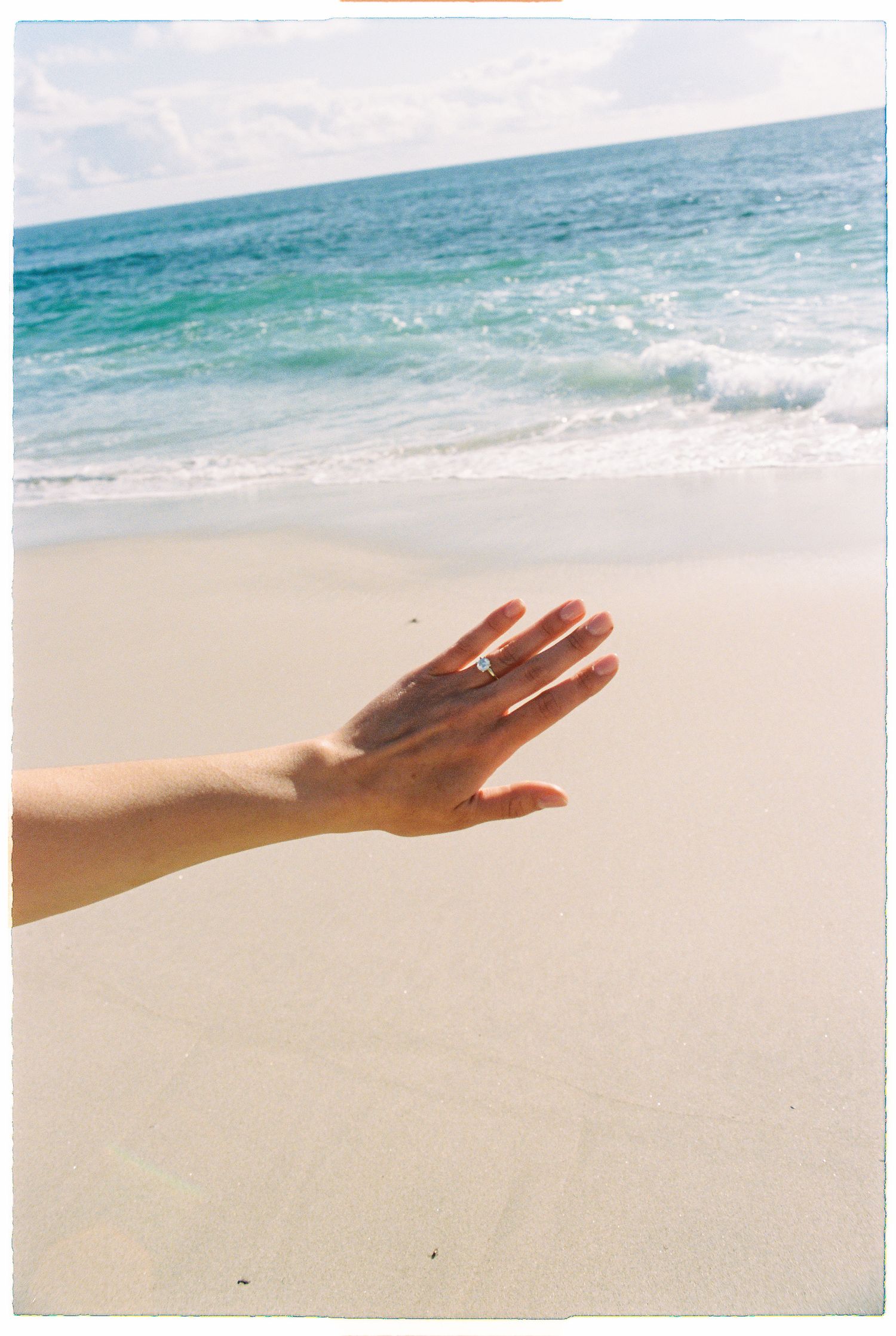 Close-up of hand with engagement ring outstretched toward turquoise ocean waves on a white sandy beach.