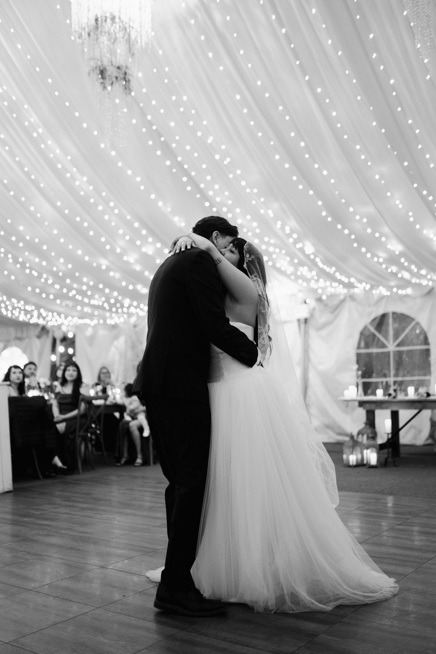 Bride and groom share first dance kiss under twinkling fairy lights at romantic wedding reception.