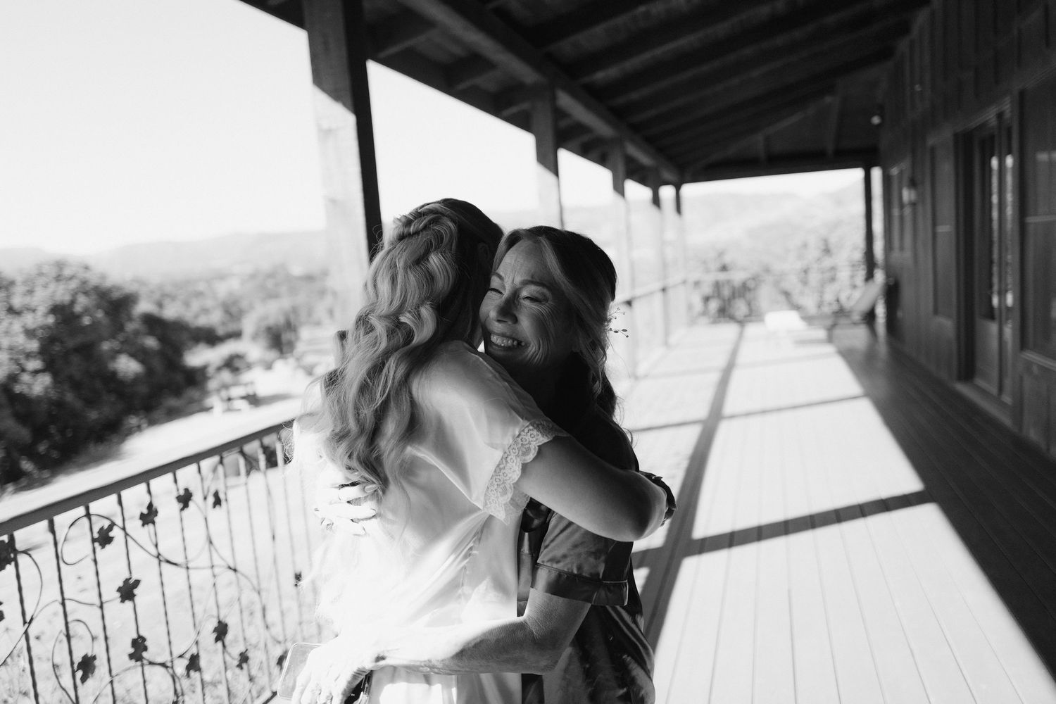 Two women sharing a warm embrace on a sunlit wooden balcony with mountain views.
