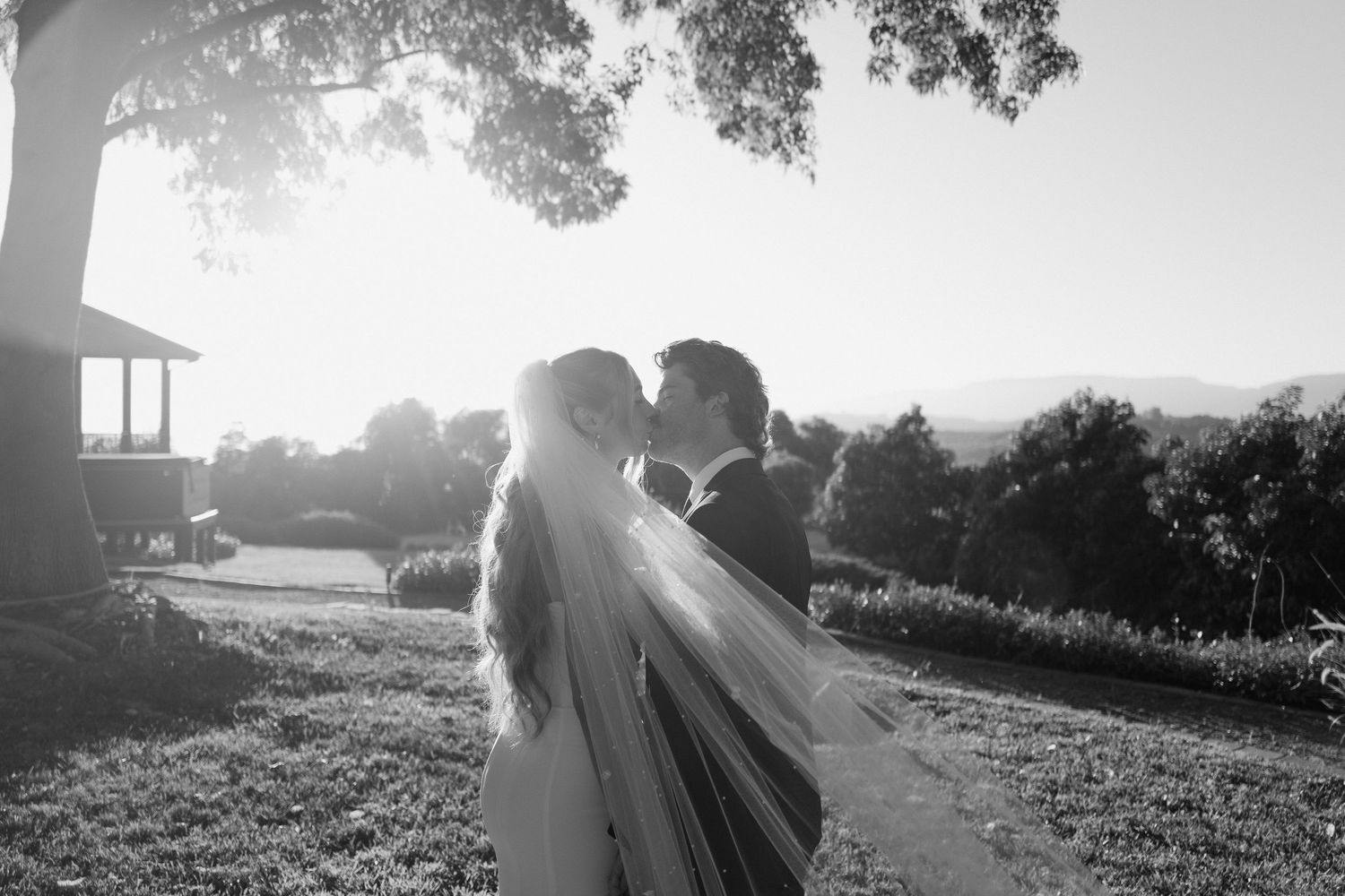 Black and white romantic photo of bride and groom kissing at sunset with long veil blowing gently.