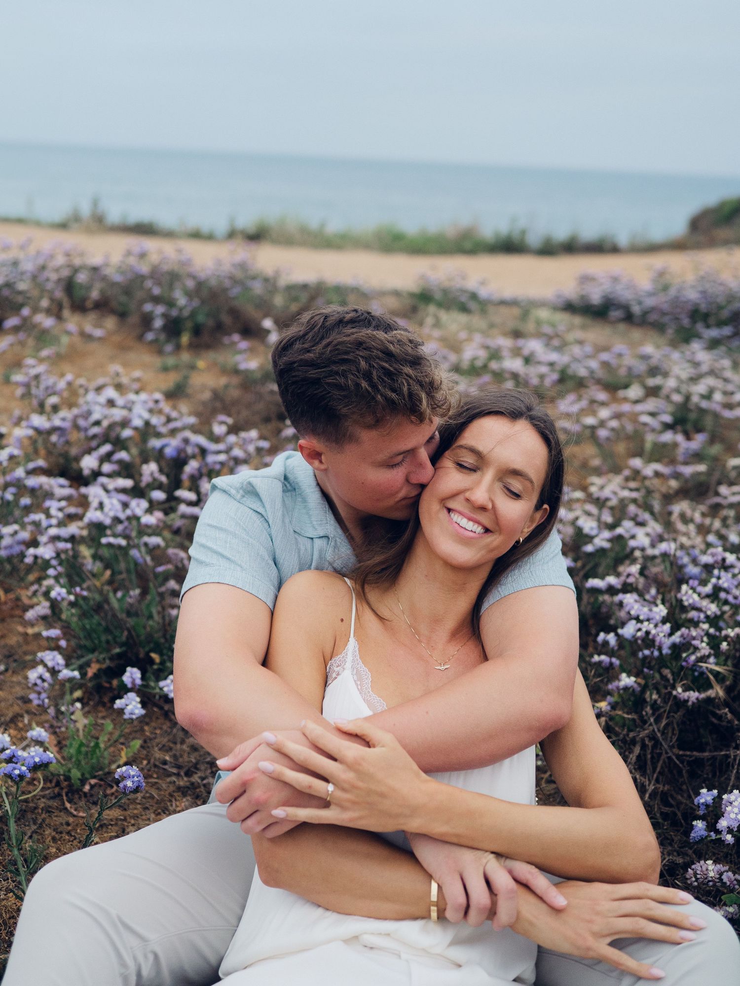 Happy couple sitting together in a field of purple wildflowers near the ocean coastline.