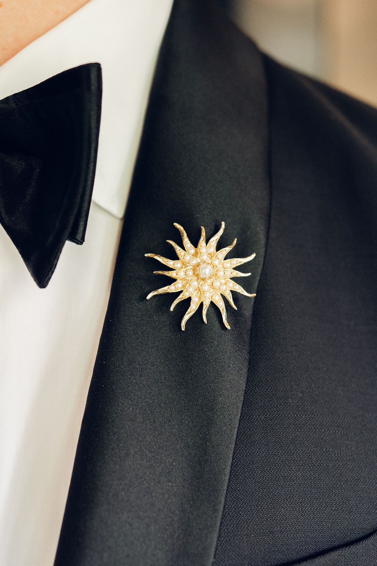 Close-up of gold sunburst brooch with pearl center pinned to black tuxedo lapel with white shirt and bow tie.