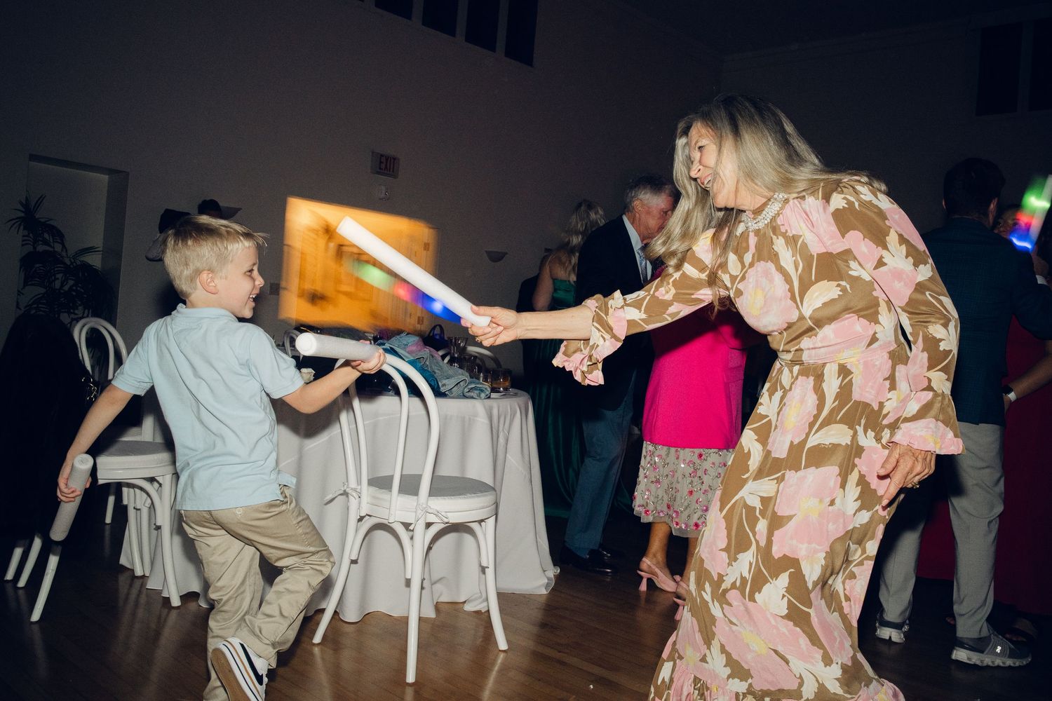 A young boy playfully duels with a glowing light-up sword with a woman in a floral dress at a wedding reception.