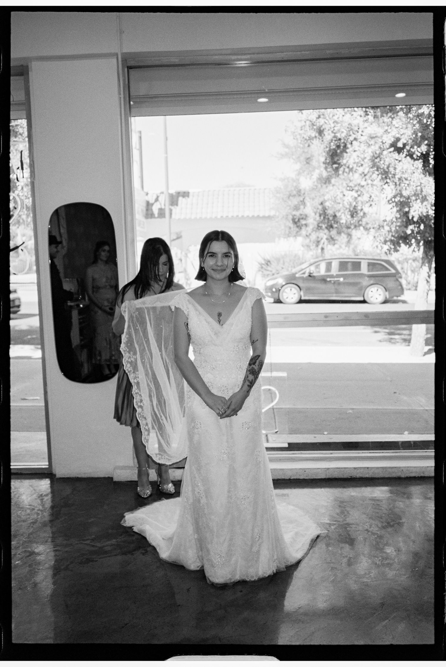 Bride in white lace wedding gown stands in bridal shop doorway with veil, black and white film photo.