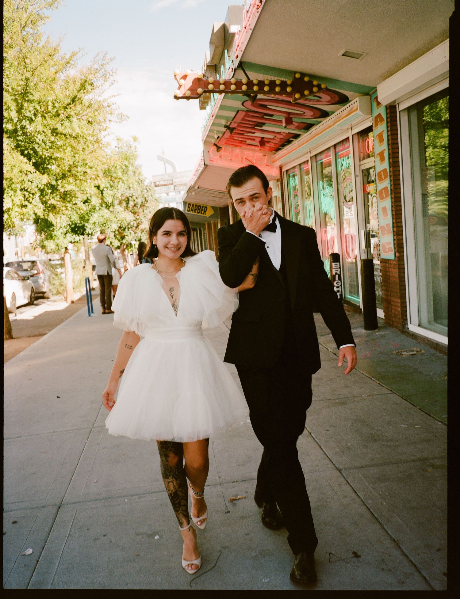 Couple in wedding attire walking on a sunny sidewalk, woman in white mini dress and man in black tuxedo.