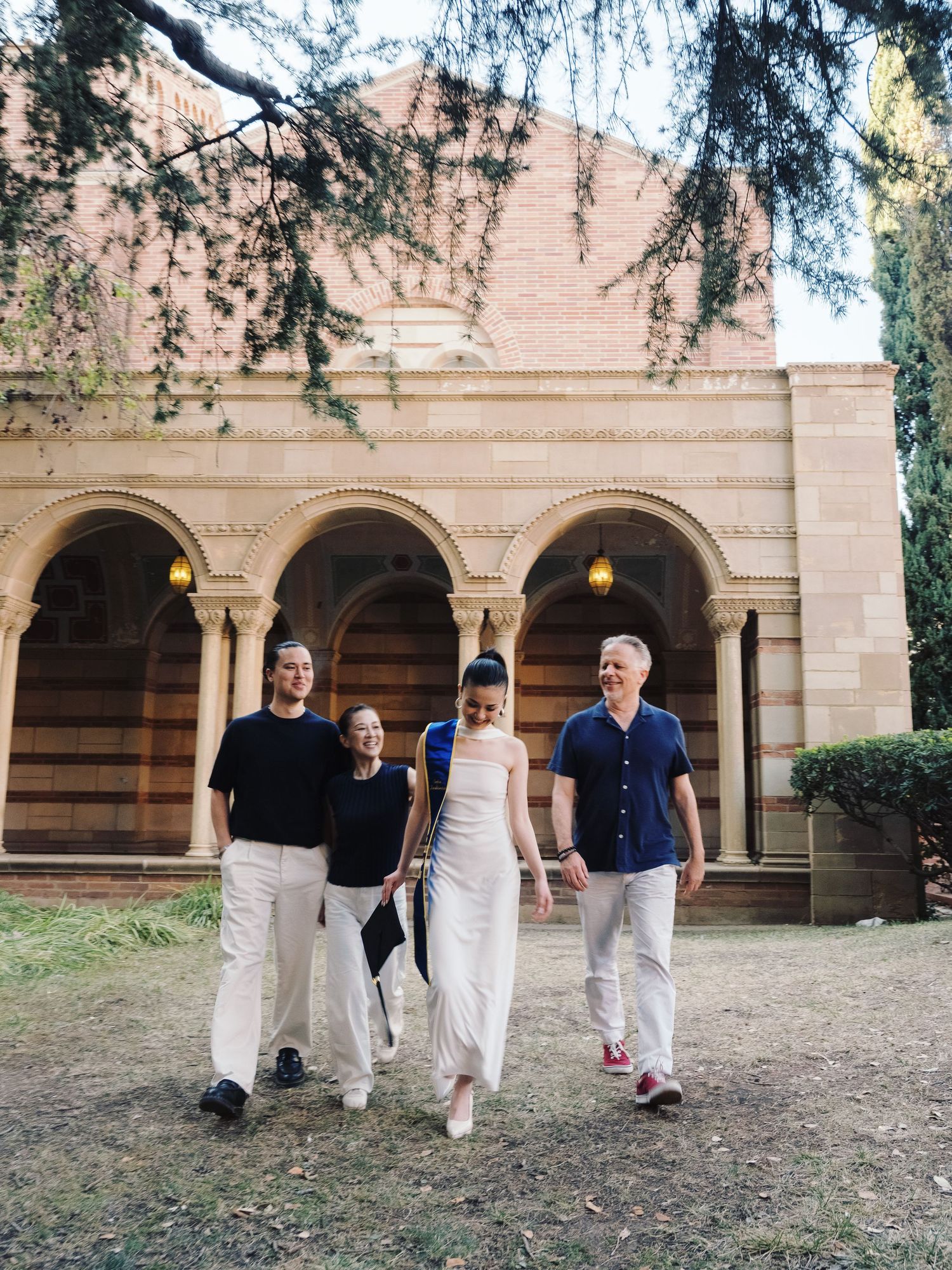 Graduate in white jumpsuit walks with family through arched courtyard of historic university building.