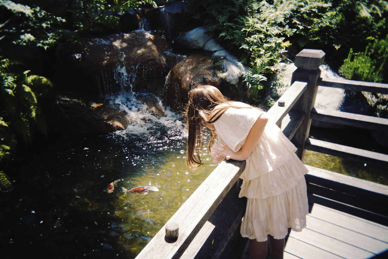 Young woman in white dress leans over wooden bridge railing to observe koi fish in garden pond.