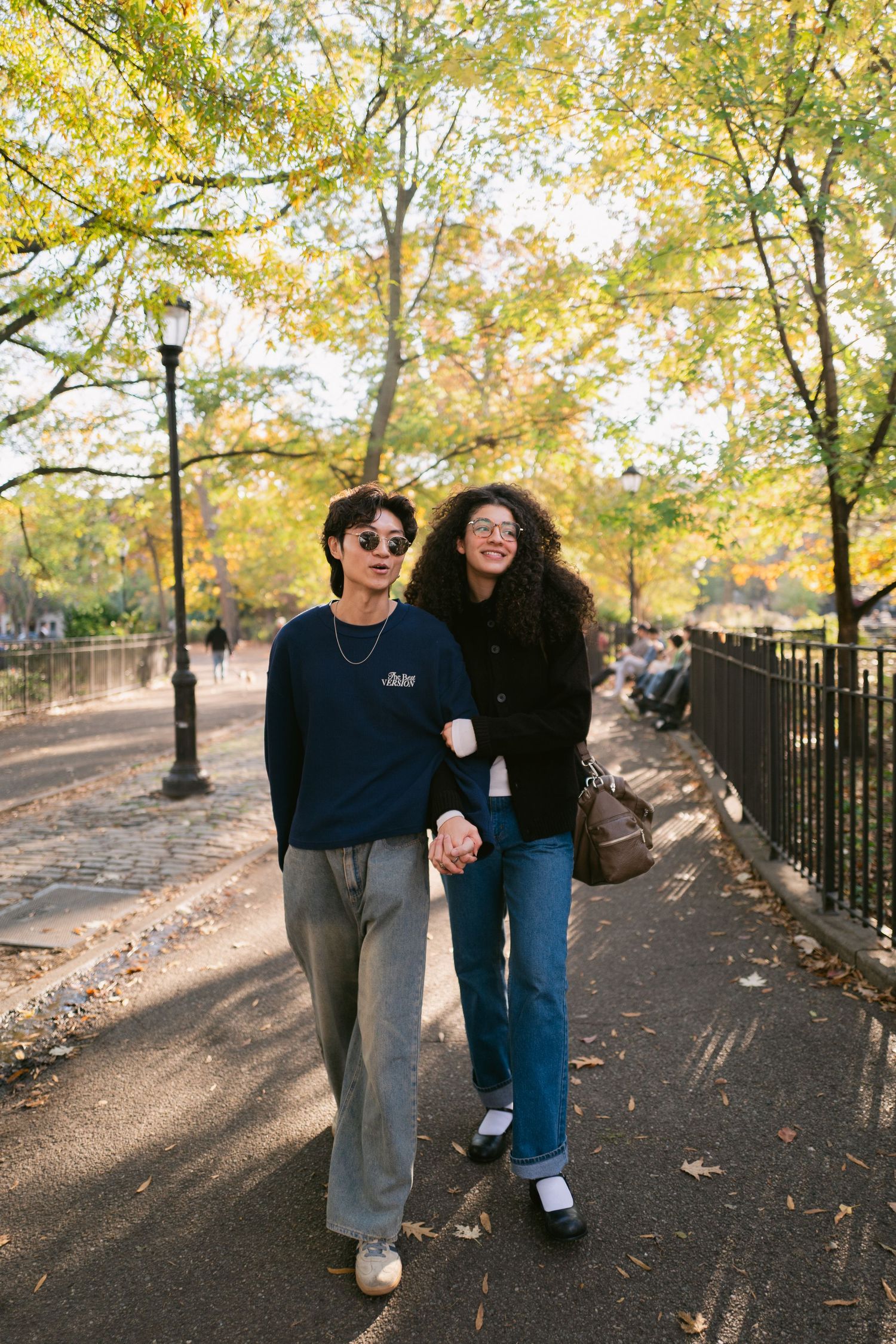 Young couple holding hands and walking together along a tree-lined park path in autumn.